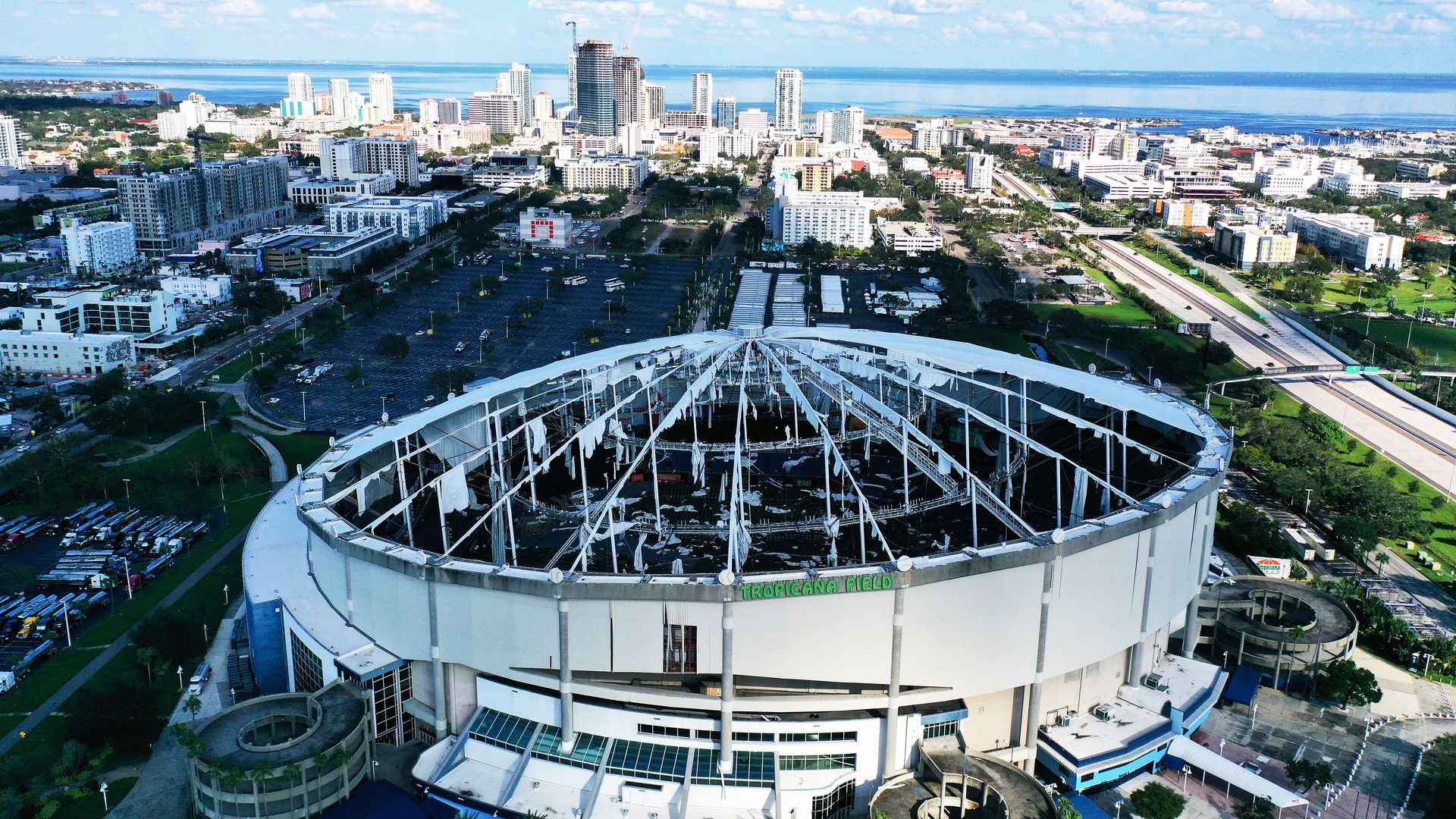 Aerial view of Tropicana Field stadium with partially collapsed roof in a cityscape under a blue sky with scattered clouds and a body of water in the background.