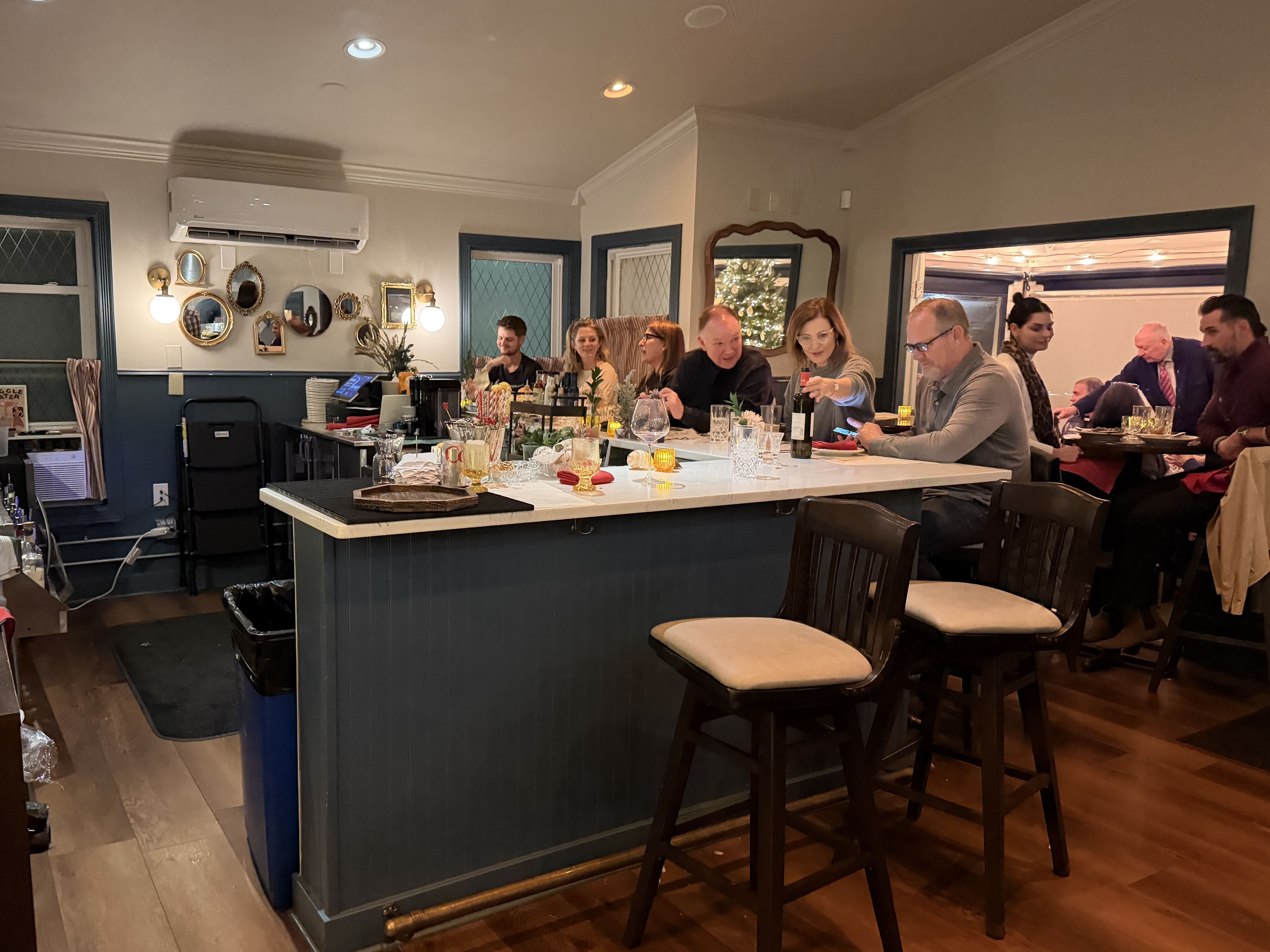 Interior of a cozy bar with people seated at and around a white marble counter with drinks. Warm lighting, wooden floor, blue walls, decorative mirrors, and a Christmas tree reflected in a mirror.