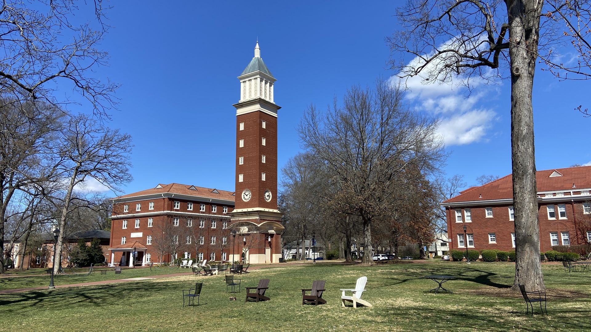 Tall red brick clock tower with white top between two red brick buildings under a clear blue sky; leafless trees and scattered chairs on green grass lawn in foreground.