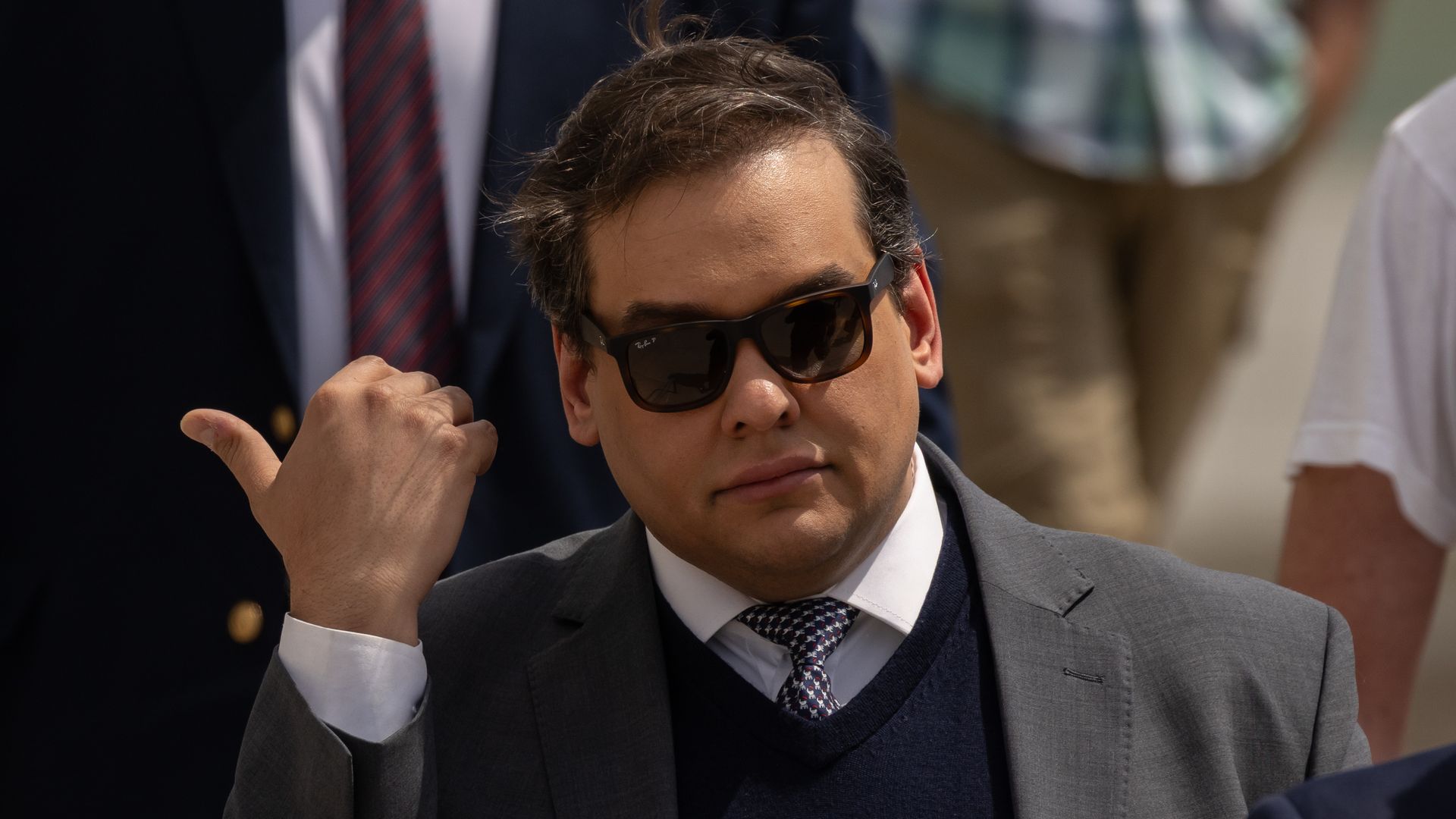 George Santos, wearing a grey suit and sunglasses, holds his hand near his face and points his thumb to the side.