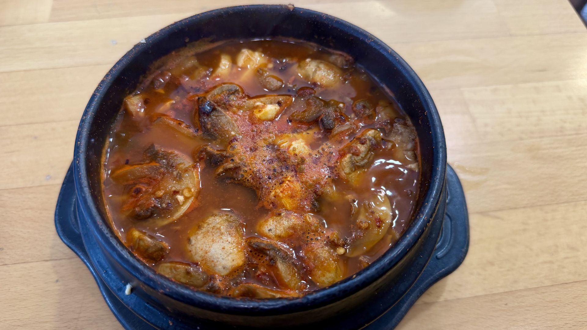 A black stone bowl filled with a spicy red stew containing mushrooms and tofu, placed on a wooden table with chopsticks partially visible in the background.
