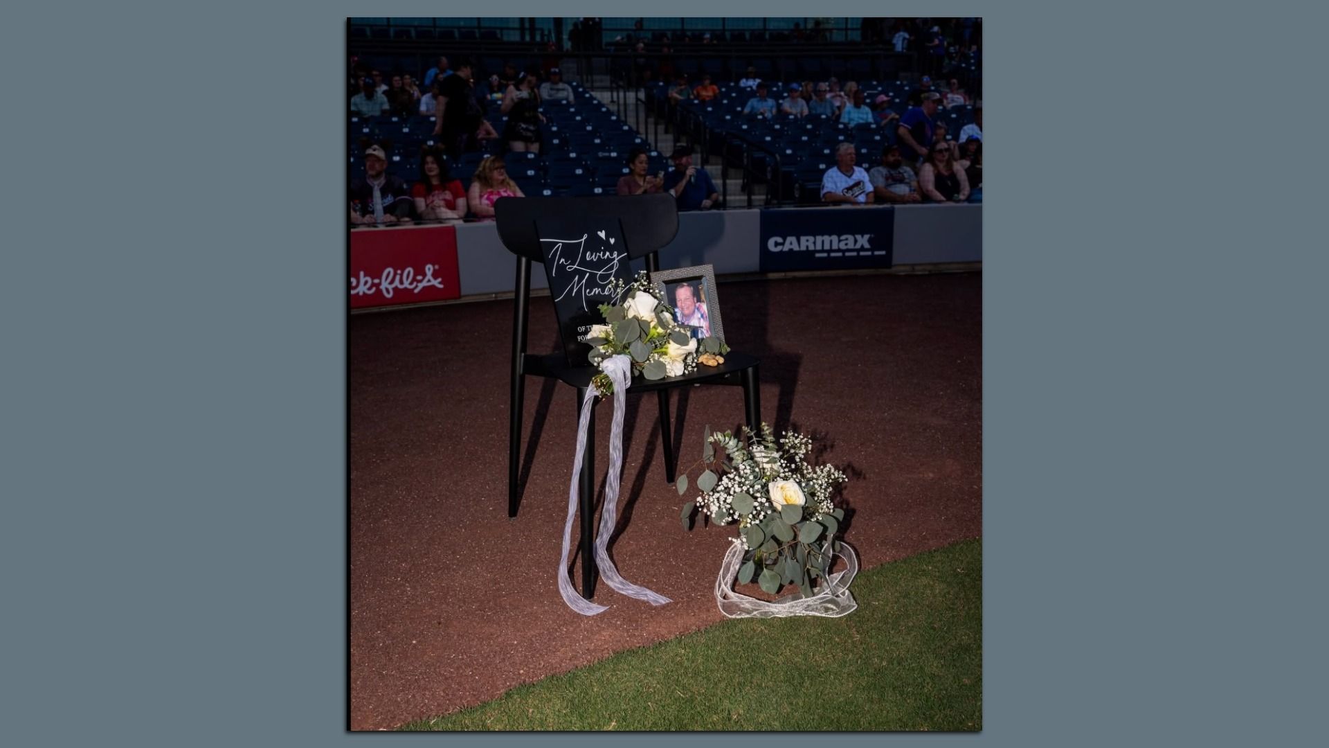 Black chair on a dirt field at a baseball stadium holds a memorial: a framed photo and a sign reading "In Loving Memory," with a white rose bouquet and ribbons, and empty blue seats in the background.