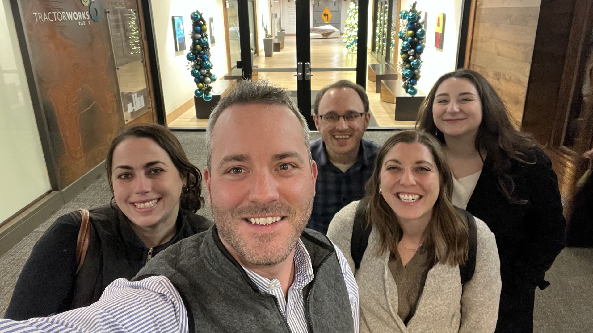 Group selfie of five smiling adults indoors near a glass door with holiday decorations including blue and silver ornaments and a large Christmas tree in the background.
