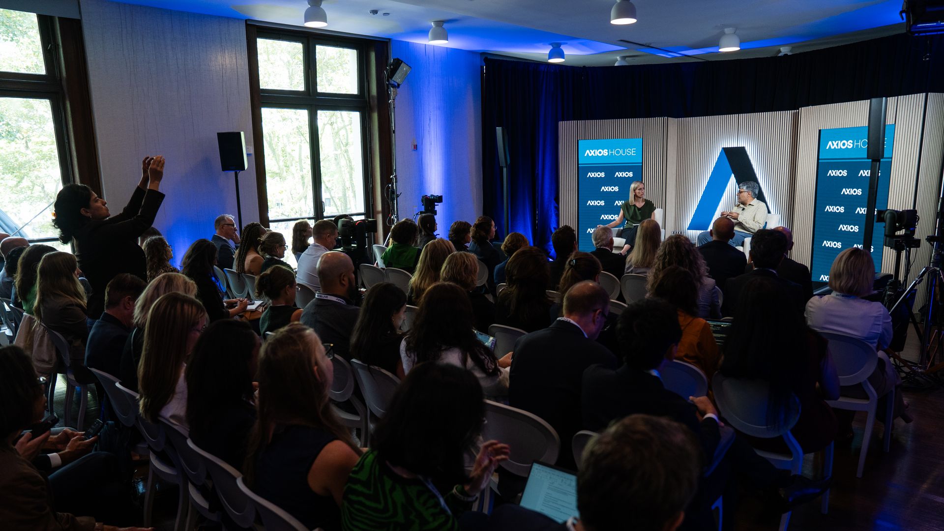Audience watches a panel on a blue-lit stage with a striped backdrop. Two speakers sit in chairs, Axios House banners beside them; a person in the foreground raises a phone to capture the talk.