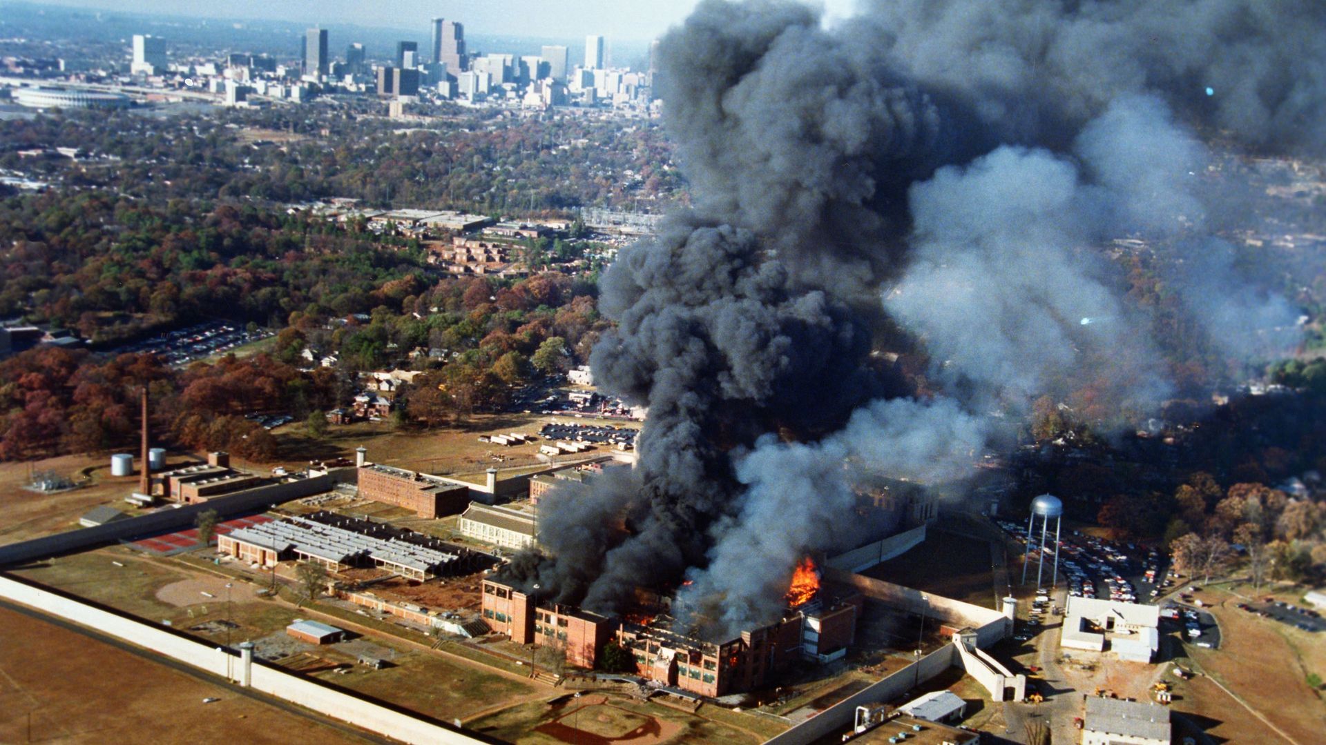A brick building on a penitentiary campus billows dark smoke from a fire with the city skyline in the background