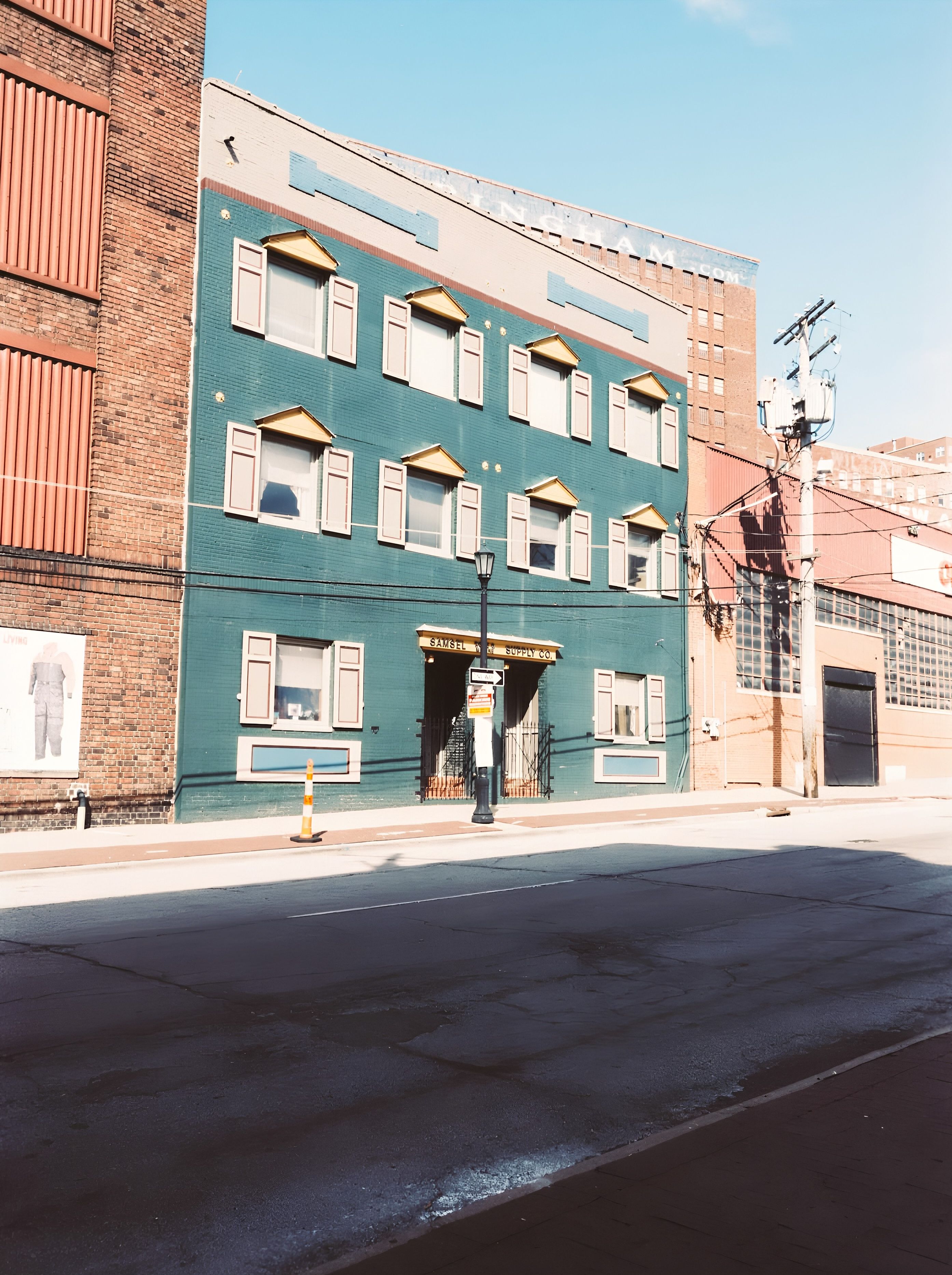 Teal brick building with three stories of white-trimmed windows and yellow triangular awnings, flanked by brick structures. Clear blue sky, street lamp, power lines, and an orange traffic cone in front.