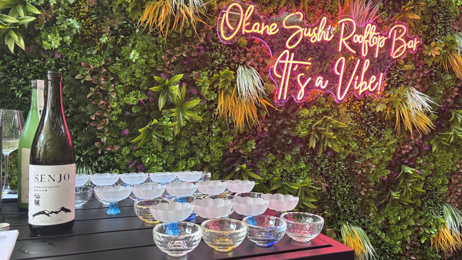 Bright pink neon sign on a dense green plant wall reads "Rooftop Bar" with "It's a Vibe". In the foreground, wine bottles and clear glass bowls sit on a dark table.