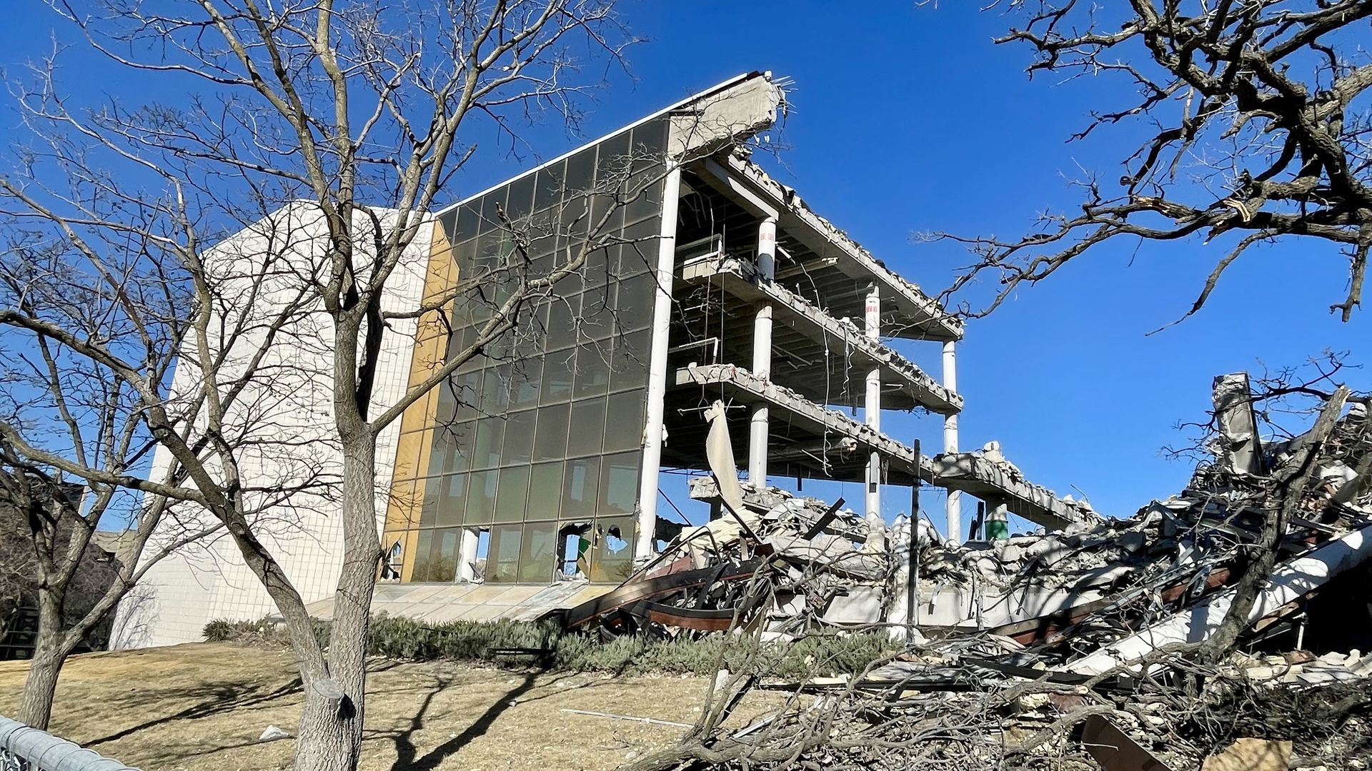Partially demolished modern building with glass facade and exposed concrete floors, surrounded by leafless trees and rubble under a clear blue sky.