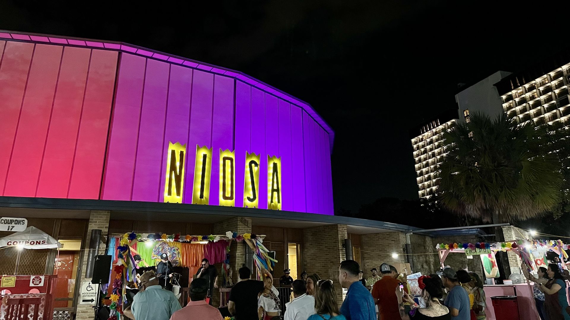 Nighttime view of a pink-purple curved building with a tall yellow sign reading NOUJA. A crowded market with stalls and festive decorations sits below, dark sky overhead and a lit apartment block to the right.