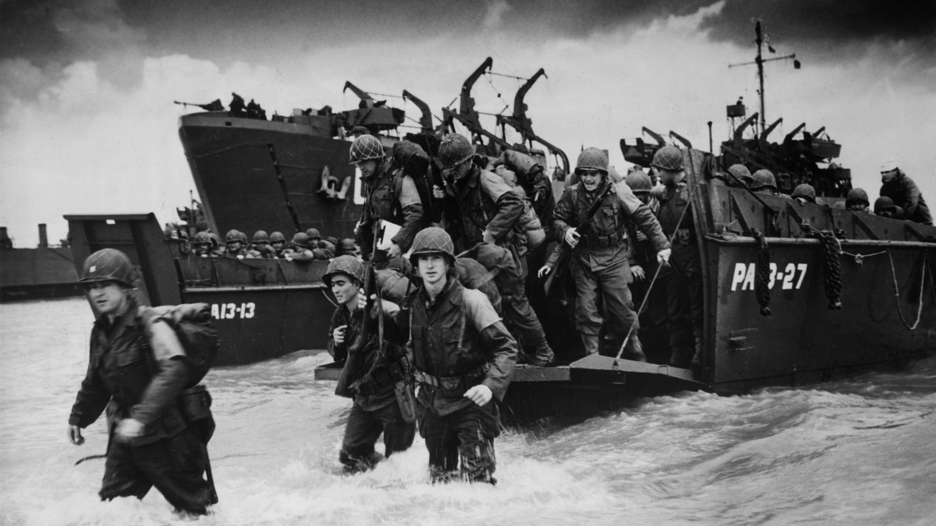 Black and white photo shows soldiers getting off a boat and into the water on D-Day.
