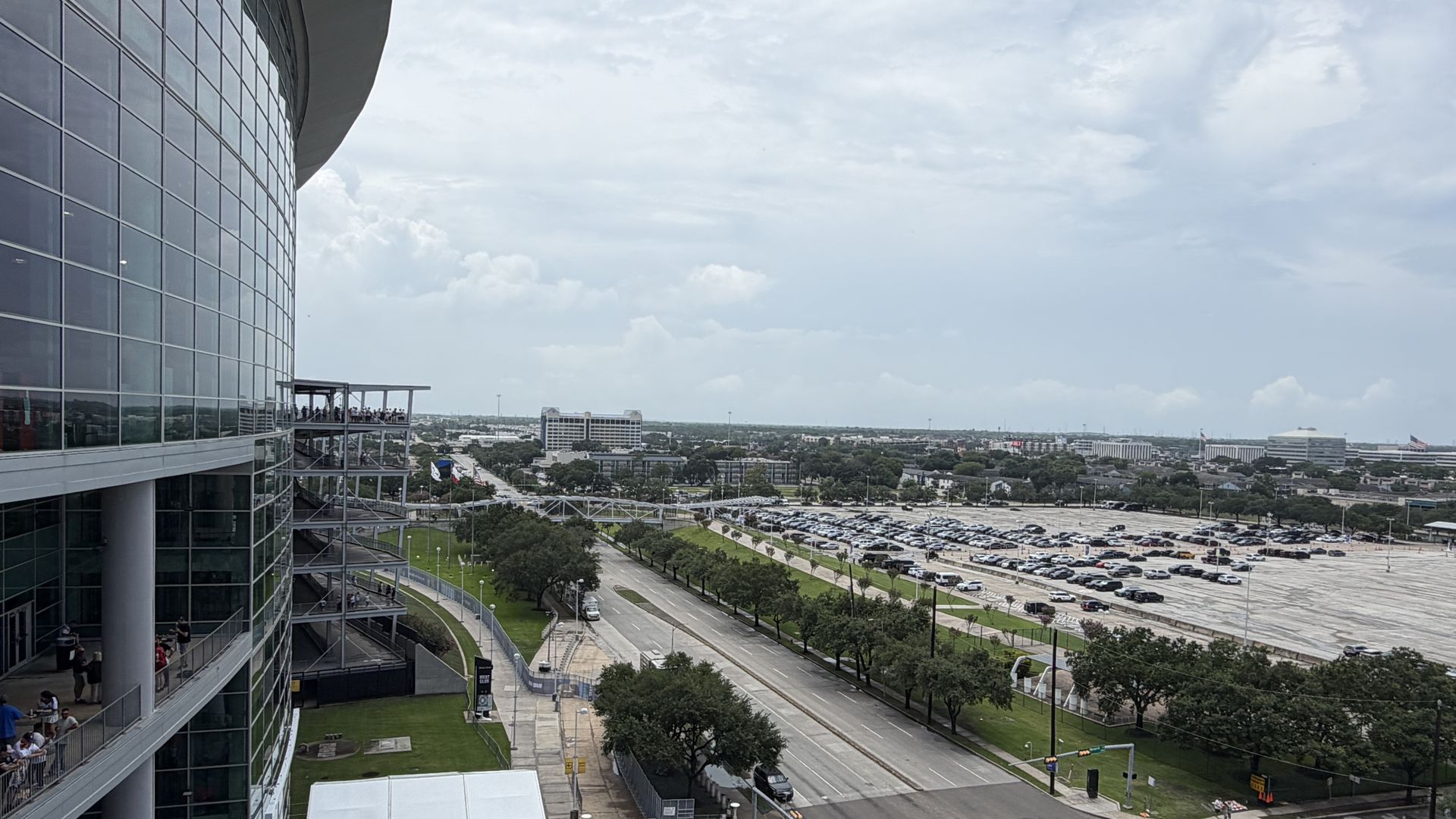 Curved glass building on the left with people on balconies, overlooking a large parking lot and distant city under a cloudy blue sky.