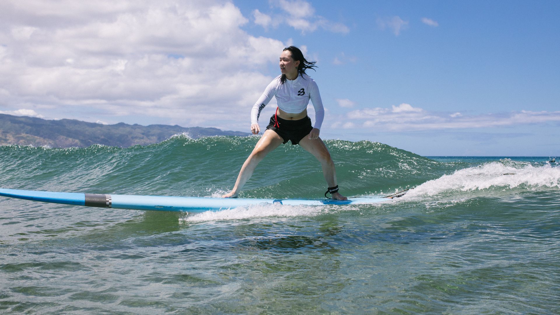 Photo of Shawna Chen standing on a surfboard and looking into the distance as she rides a wave