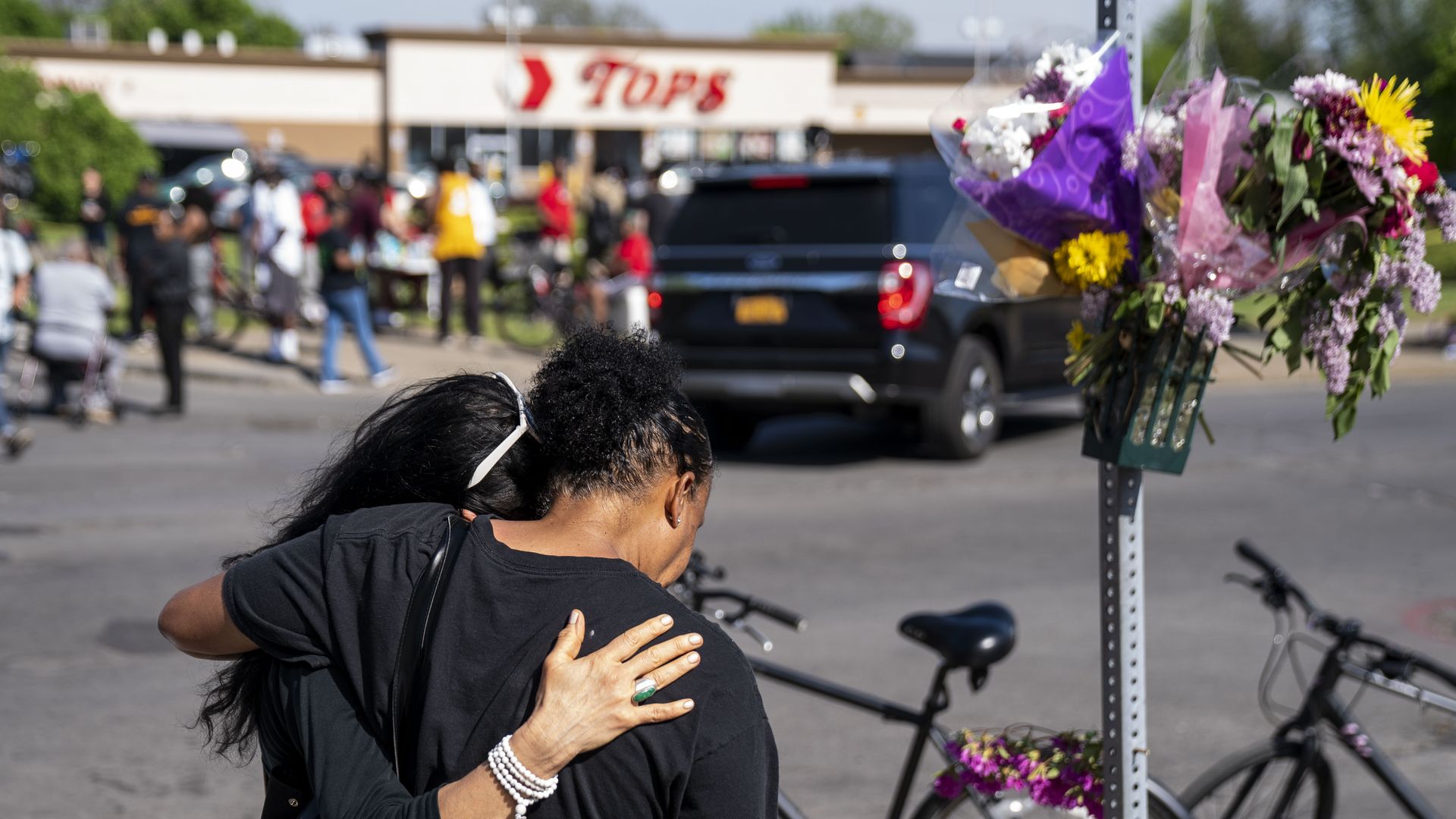 Photo of two people embracing in front of the Tops grocery store where police cars gathered in response to a shooting