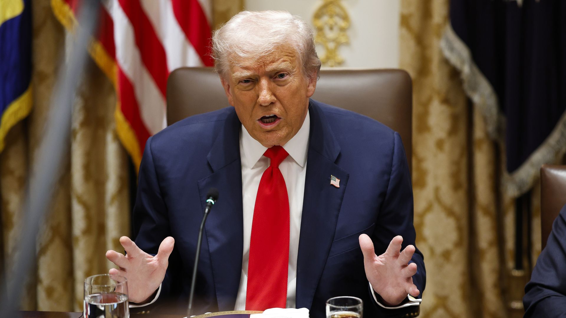 President Trump, wearing a navy jacket with a US flag pin, white shirt and red tie, gestures at a brown desk that has a glass of water and a glass of coke on it as he speaks into a black mic  in the Cabinet Room at the White House.