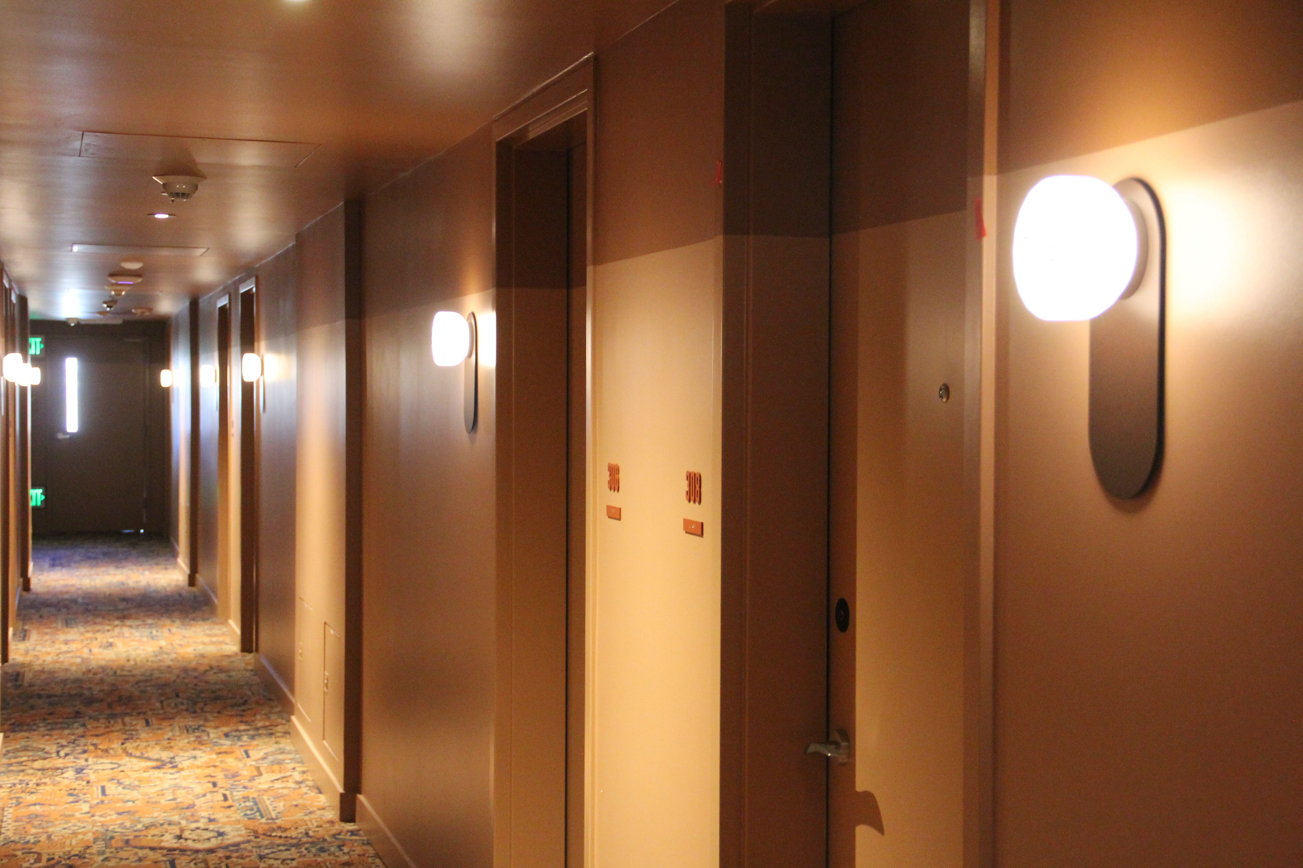Long hotel corridor, amber-lit walls with round sconces, doors on the right, a patterned carpet, and a distant exit sign at the far end.