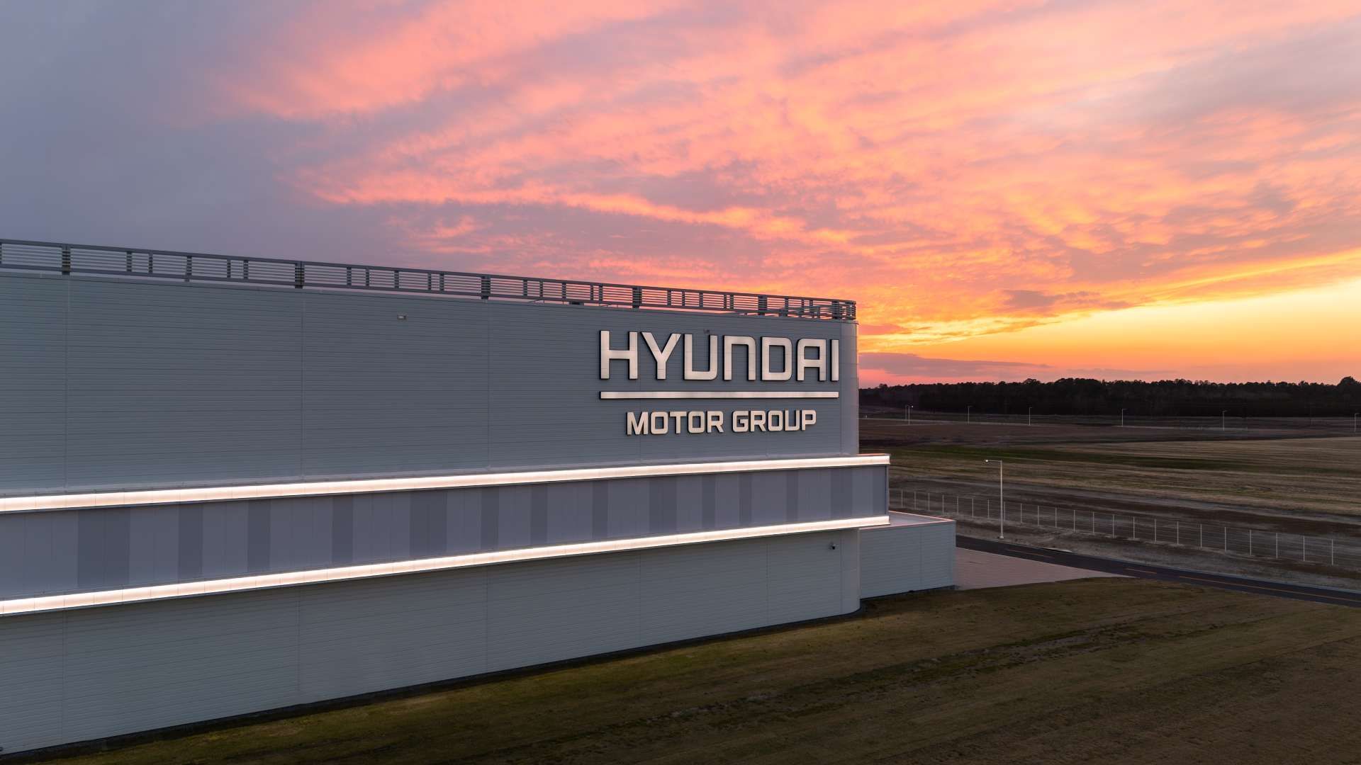 Exterior view of a gray Hyundai Motor Group building with large 3D letters on the side, illuminated stripes along the base, under a pink-orange sunset and flat fields beyond.