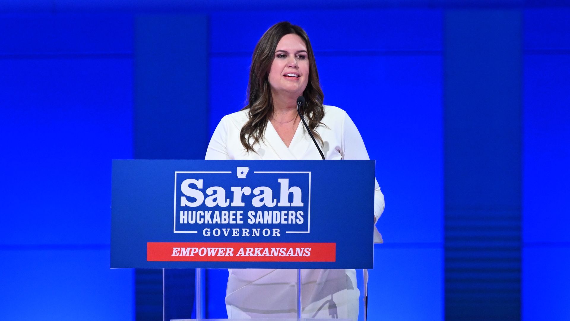 A photo of Gov. Sarah Huckabee Sanders in a white dress on a blue field giving her acceptance speech after winning the election, standing at a podium.