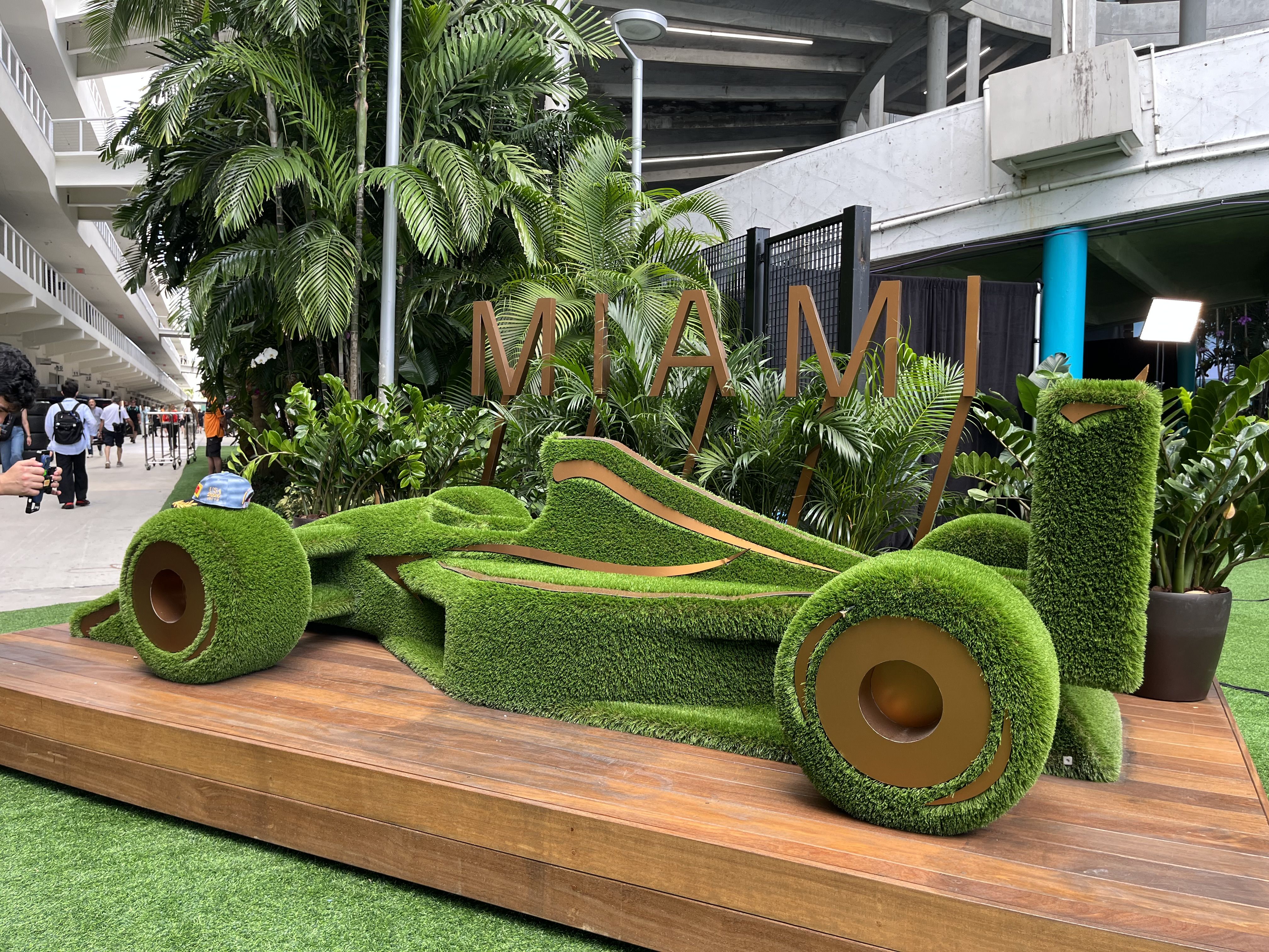 Outdoor display of large gold letters spelling MAMAMI among lush tropical plants on a wooden platform; a concrete structure and blue pillar loom in the background as people walk nearby.
