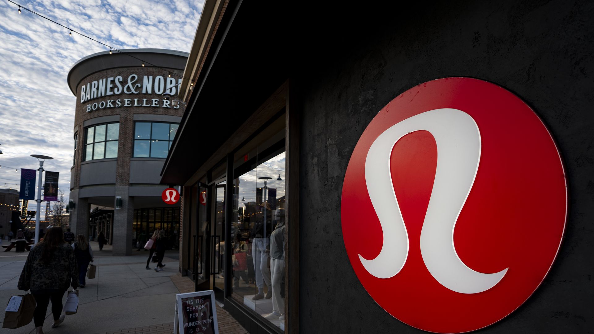 Outdoor shopping street with Barnes & Noble Booksellers on the left and a large red circular logo on a dark wall to the right; string lights overhead and shoppers with bags.