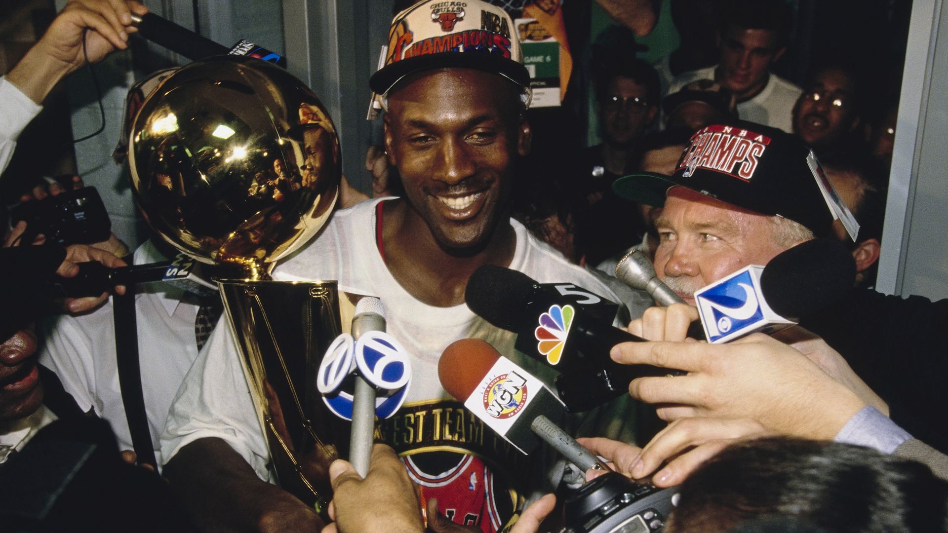 Smiling basketball player in a white jersey wearing a Chicago Bulls Champions cap, surrounded by reporters and cameras, holding a large gold trophy amid the press.