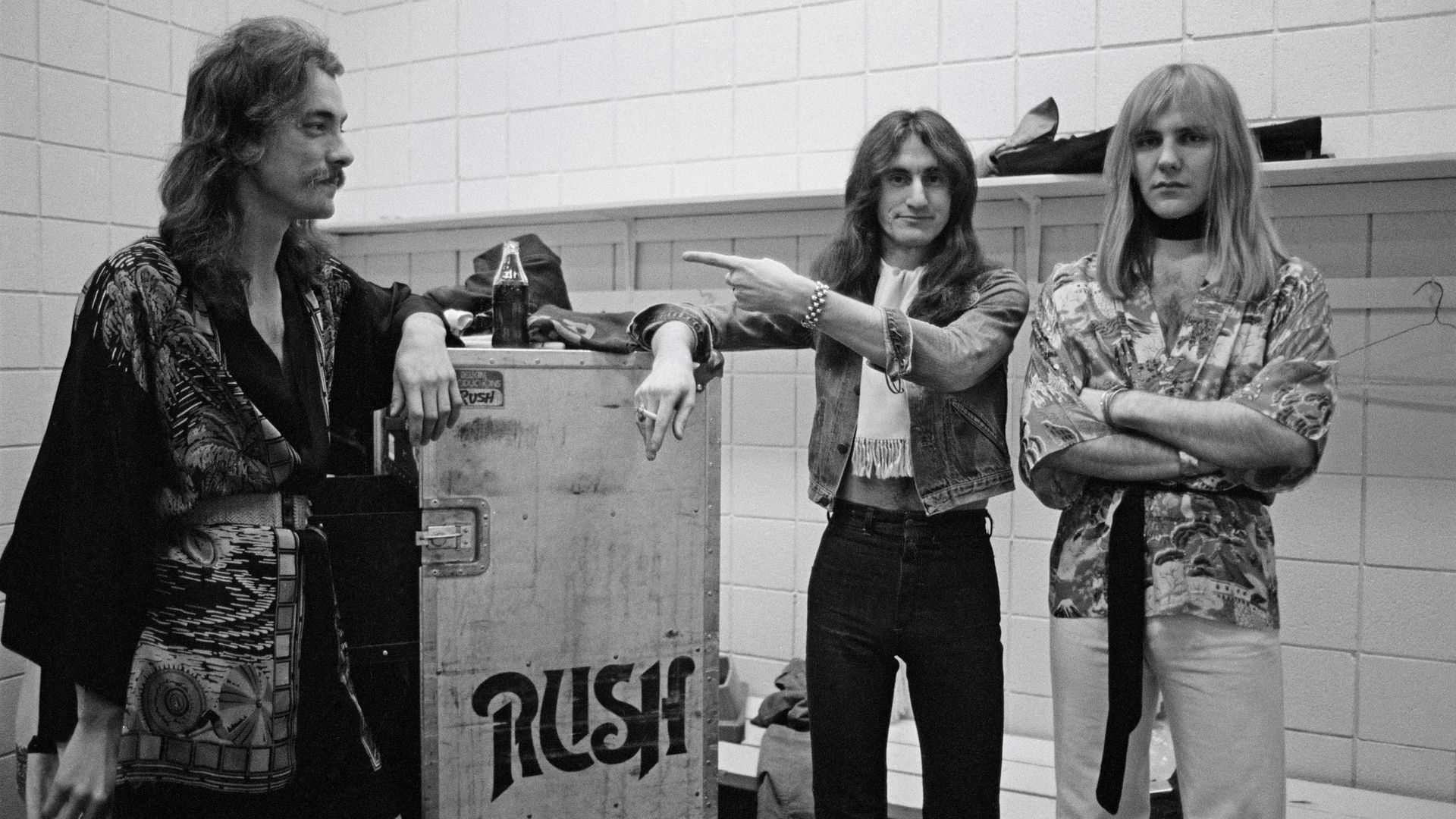 Black-and-white shot of three men backstage, leaning on a crate reading "RUSH". Left: mustached man with long hair in a patterned coat. Middle: denim-clad man points. Right: blonde in floral shirt and black turtleneck.
