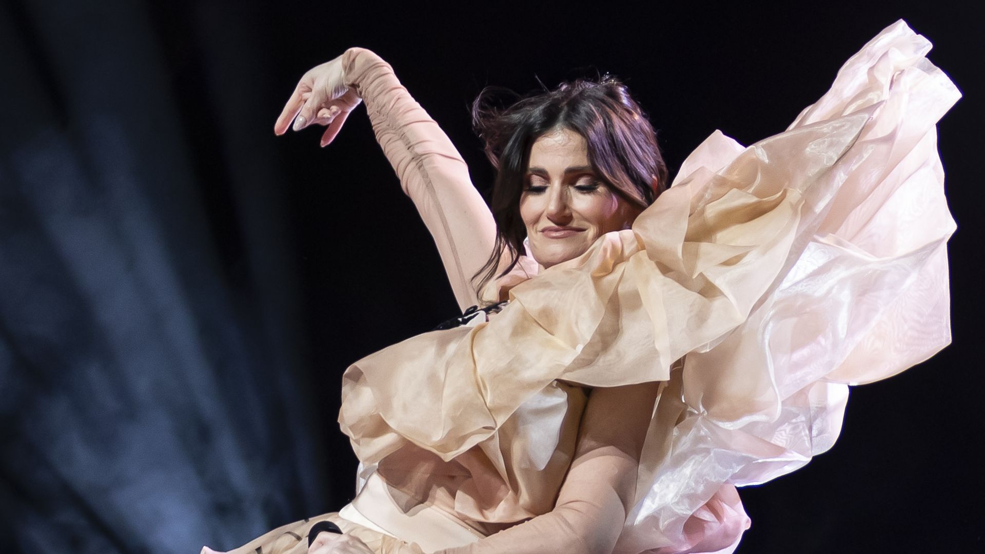 Female performer in a light peach, billowing ruffled gown, her eyes closed in a smile as she raises one arm and holds a microphone on a dark stage.