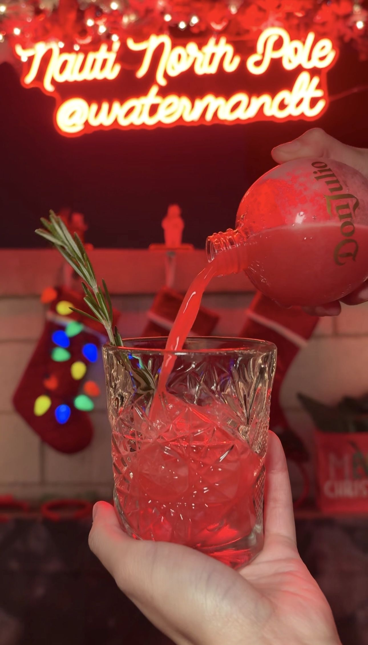 A hand pours a bright red drink from a Don Julio bottle into a glass with ice and a rosemary sprig, with red Christmas stockings and a neon sign reading "Nauti North Pole @watermandt" in the background.