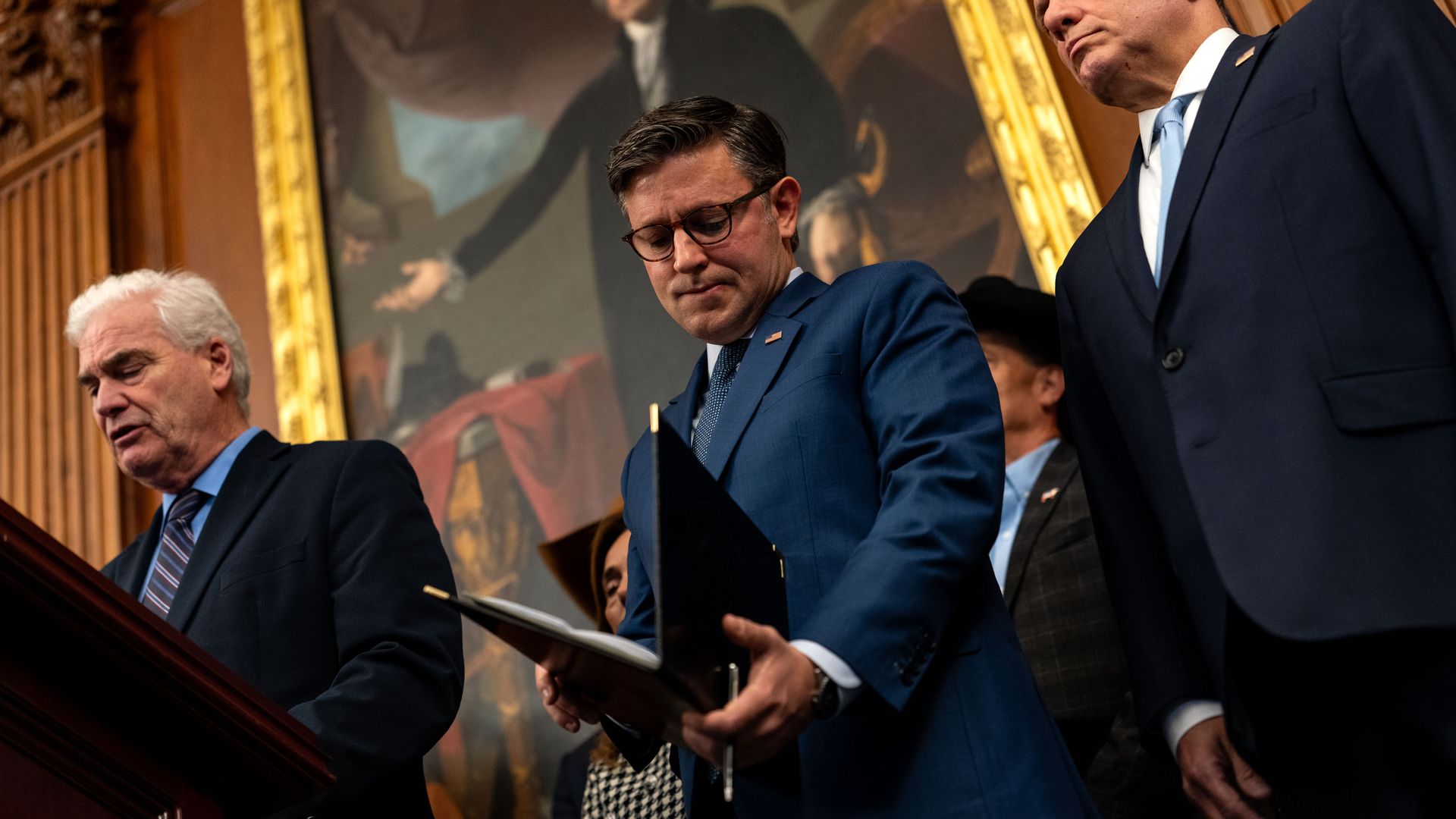 Mike Johnson wearing a blue suit and looking down at a black folder, flanked by fellow members of House Republican leadership while standing in front of a wooden wall with a large portrait of George Washington on it.