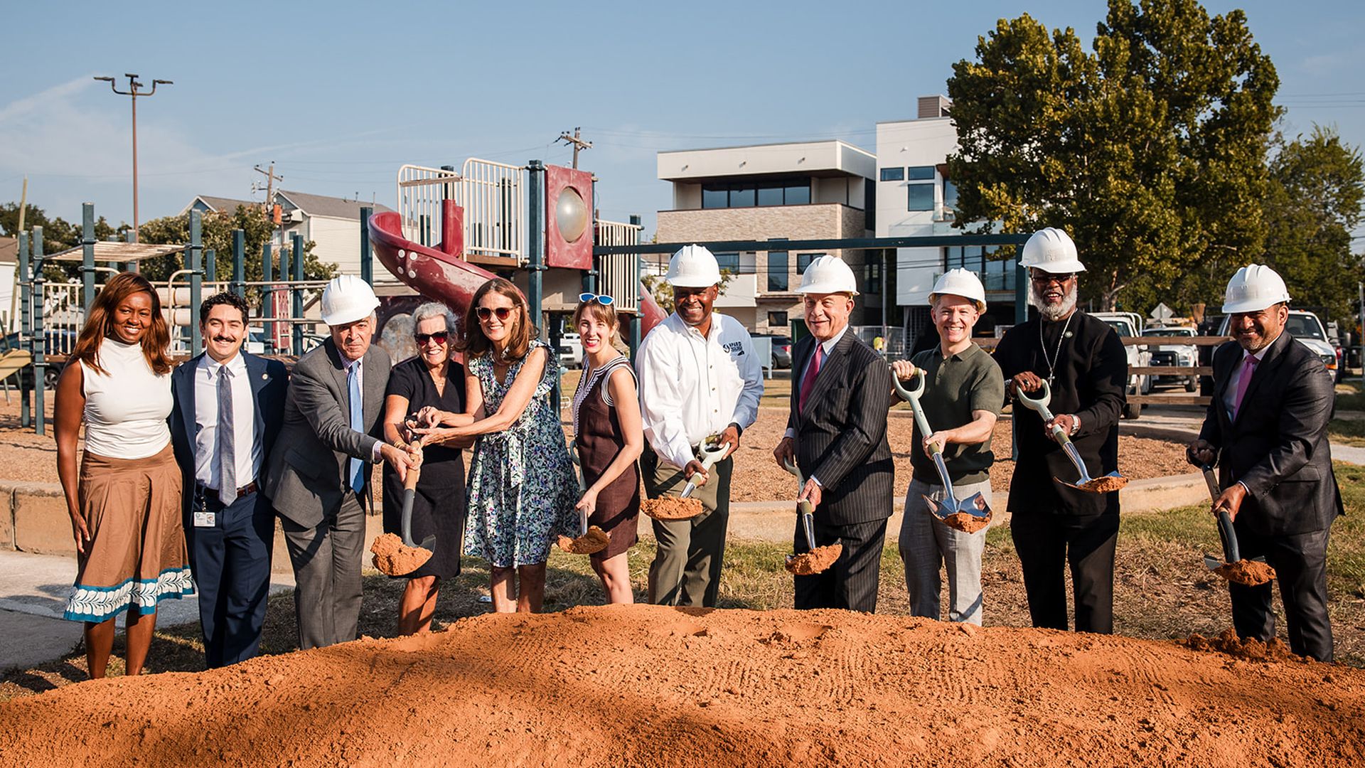Diverse group wearing hard hats and business attire shovel dirt at a playground groundbreaking ceremony; a dirt mound fills the foreground with playground equipment and buildings behind.
