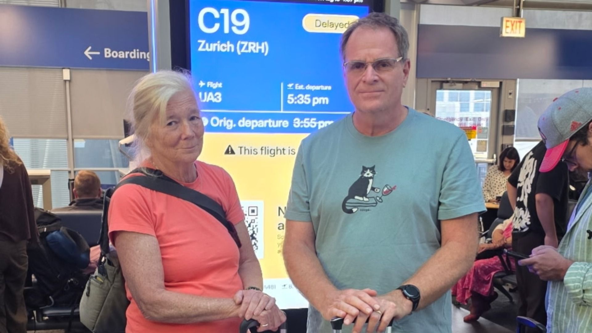 Two adults stand in an airport beside a blue flight board reading "C19 Zurich (ZRH)" and "Delayed". Woman in coral left, man in teal shirt with a cat graphic right; background travelers.