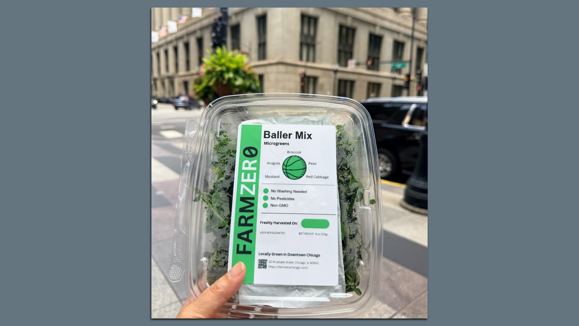 Plastic container of microgreens labeled 'Baller Mix' with a green 'FARM ZERO' label; lists broccoli, arugula, peas, mustard, red cabbage; urban street scene in background.