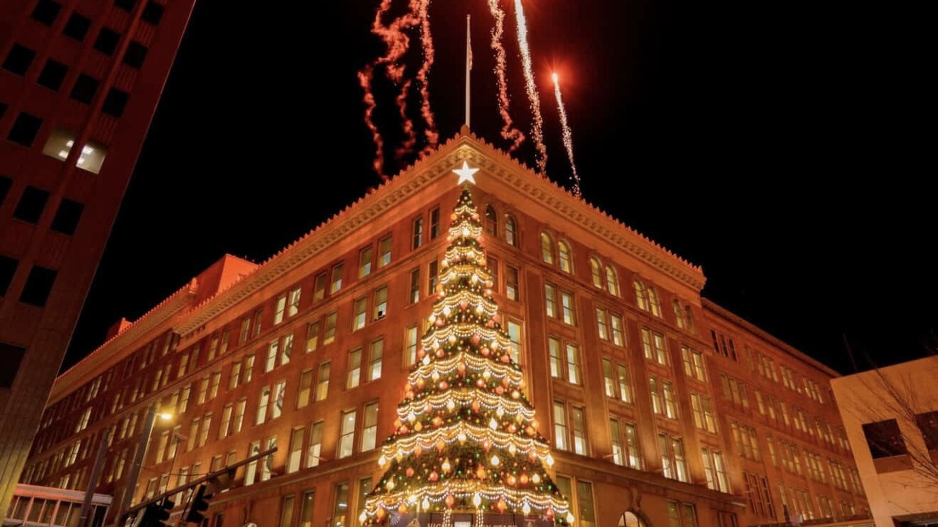Large decorated Christmas tree with lights and ornaments in front of a tall, well-lit building at night; fireworks burst in the dark sky above the building's corner.