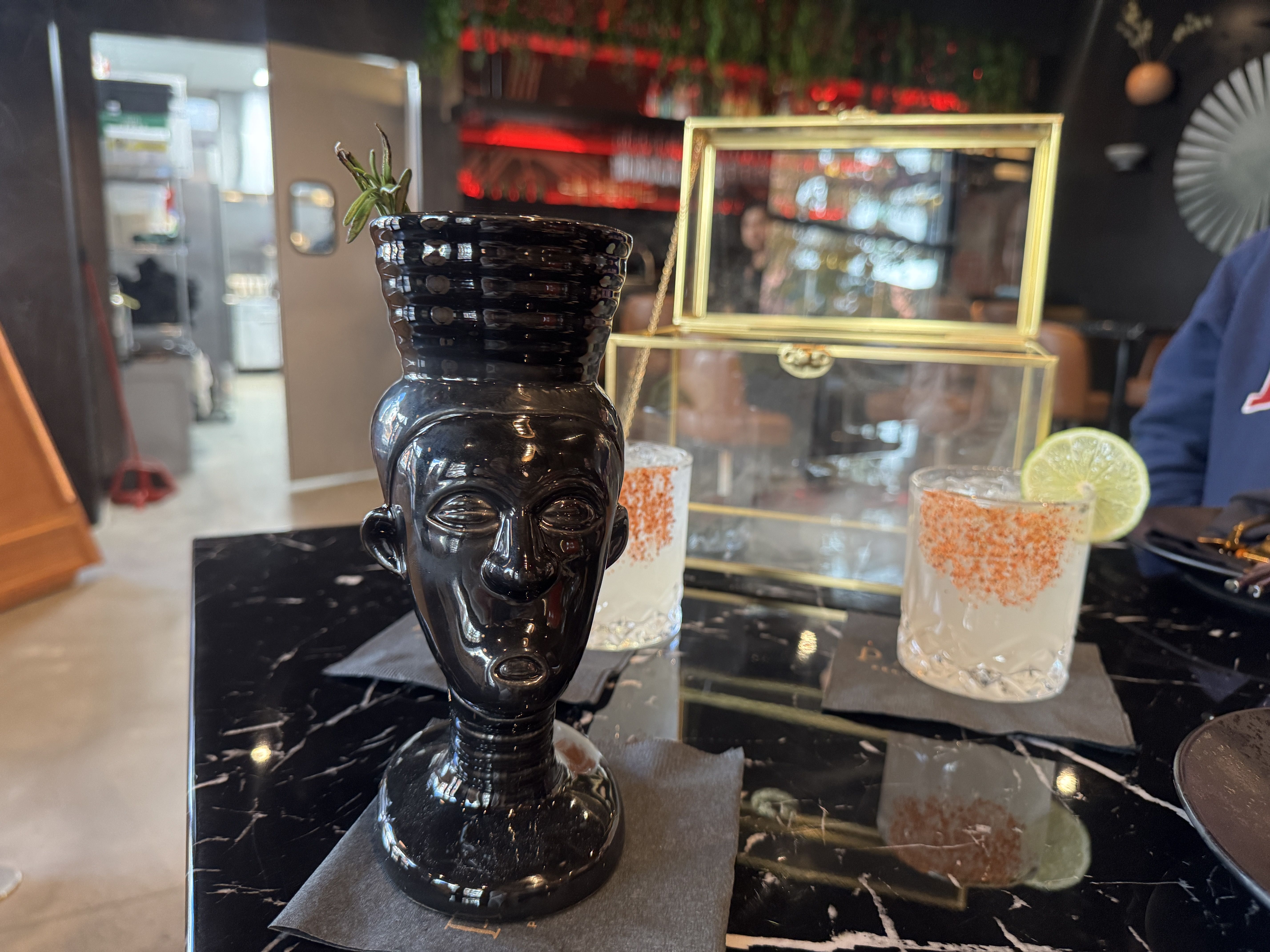 Black ceramic cup shaped like a stylized human face with rosemary garnish, sitting on a black marble table. Two clear glasses with orange-rimmed edges and lime wedges are in the background.