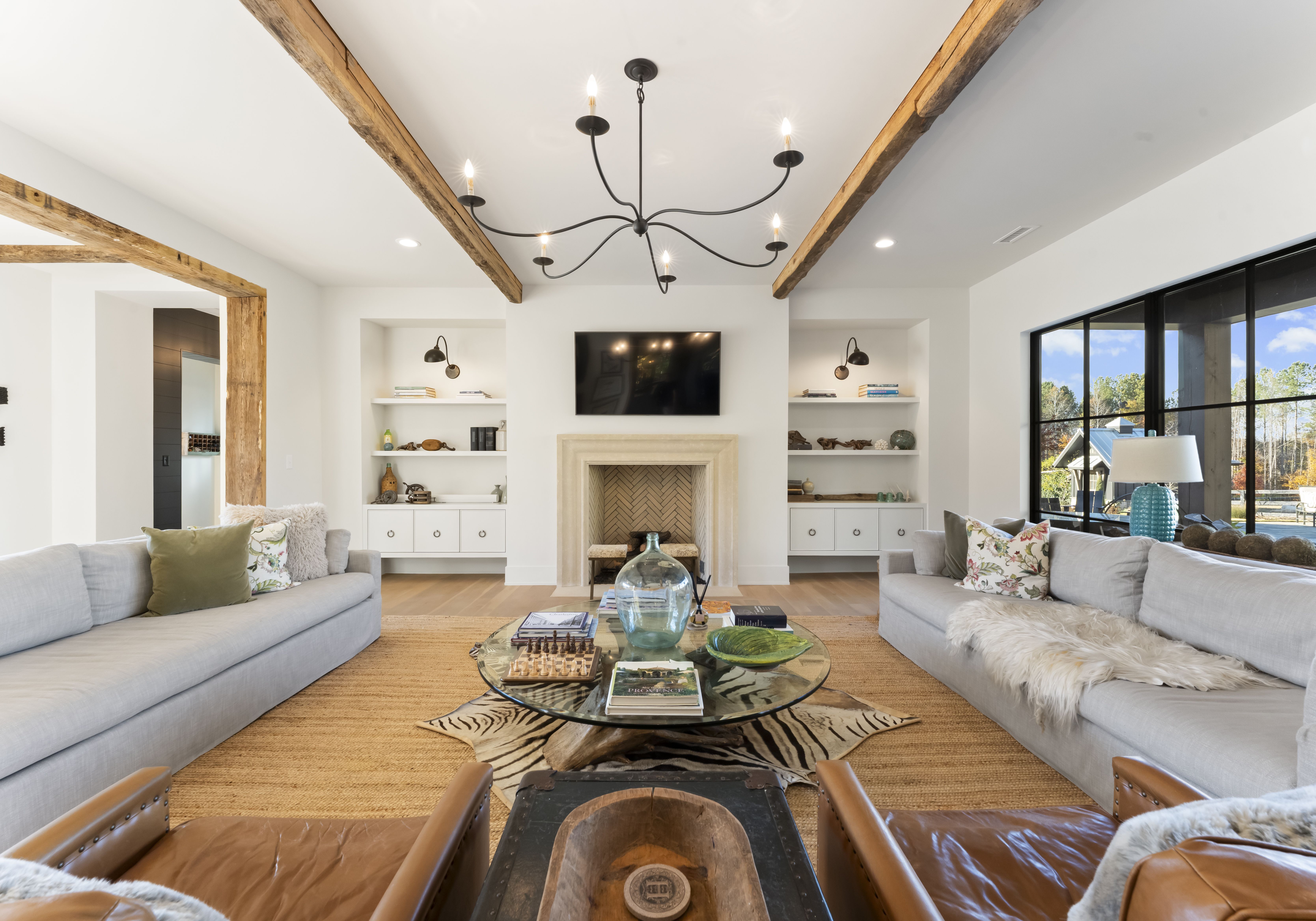 Bright living room with two light-gray sofas around a round glass coffee table on a natural jute rug. Beige fireplace with TV above, shelves, exposed wooden beams, and a black chandelier overhead.