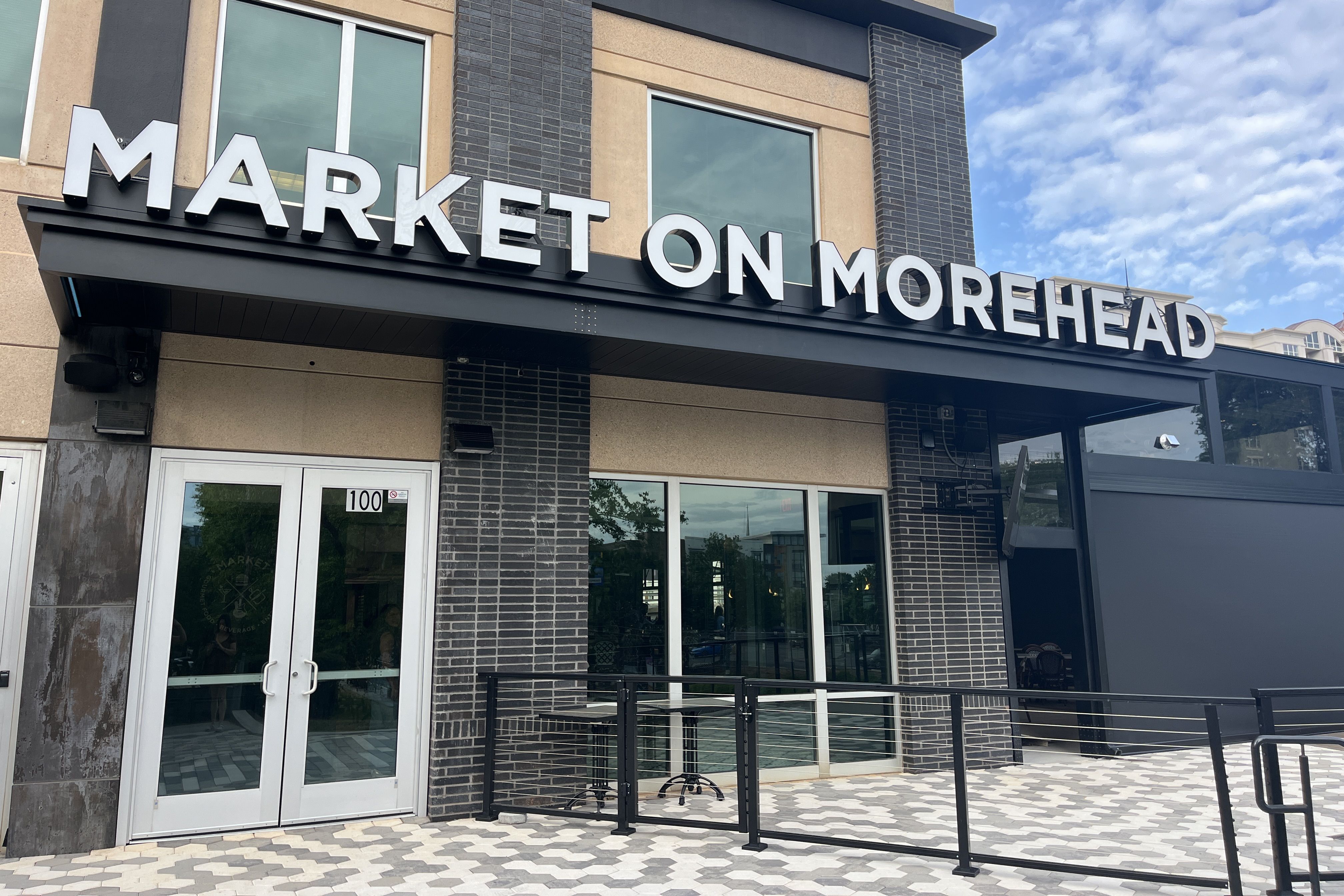 Exterior of a modern storefront with large white letters reading "MARKET ON MOREHEAD" above glass doors. Beige and dark brick facade, black railing, and a blue sky with scattered clouds.