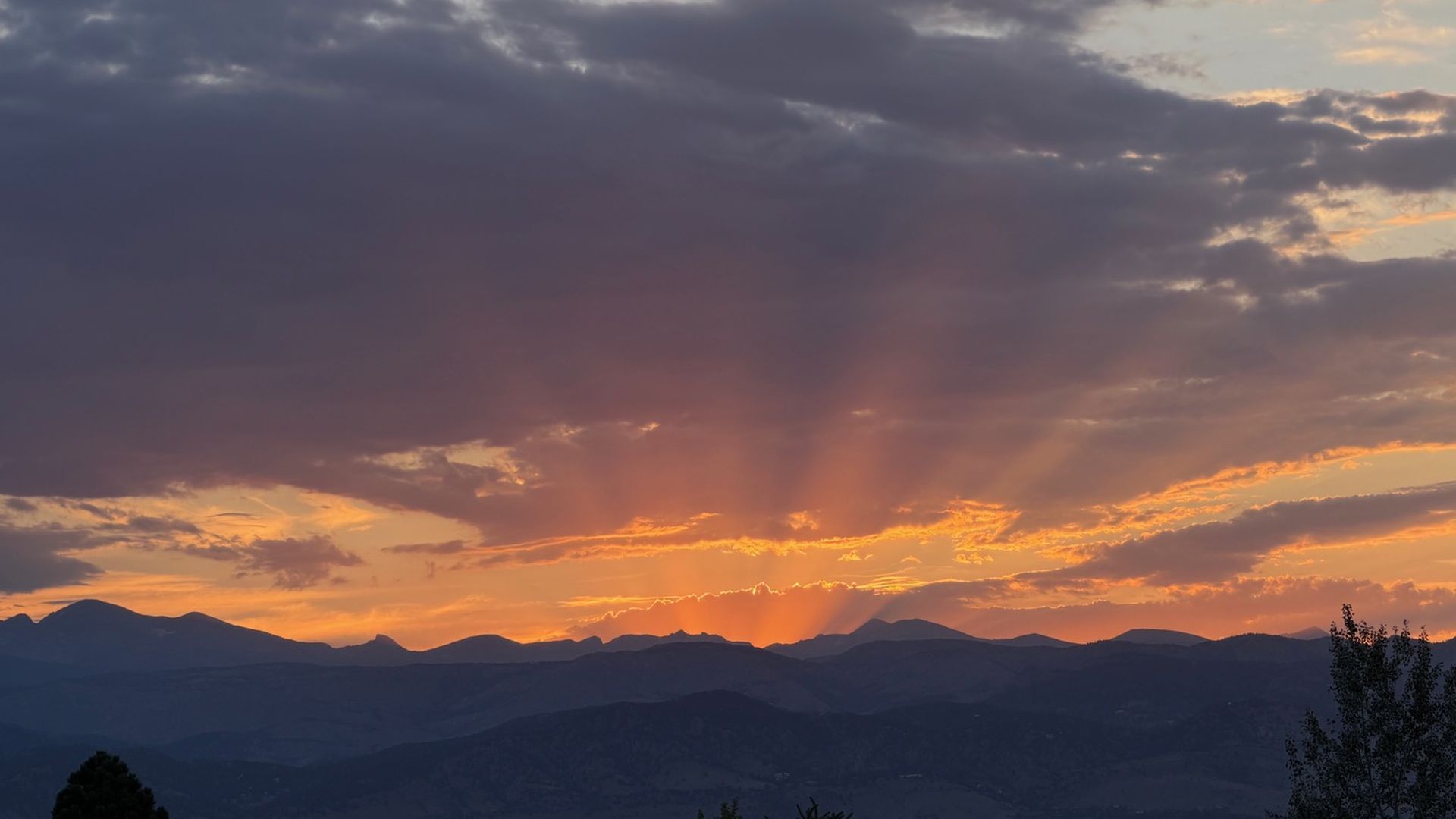 Sunset over a distant mountain range with orange light spilling through dark clouds. Sun rays fan across the horizon while silhouetted trees frame the foreground.