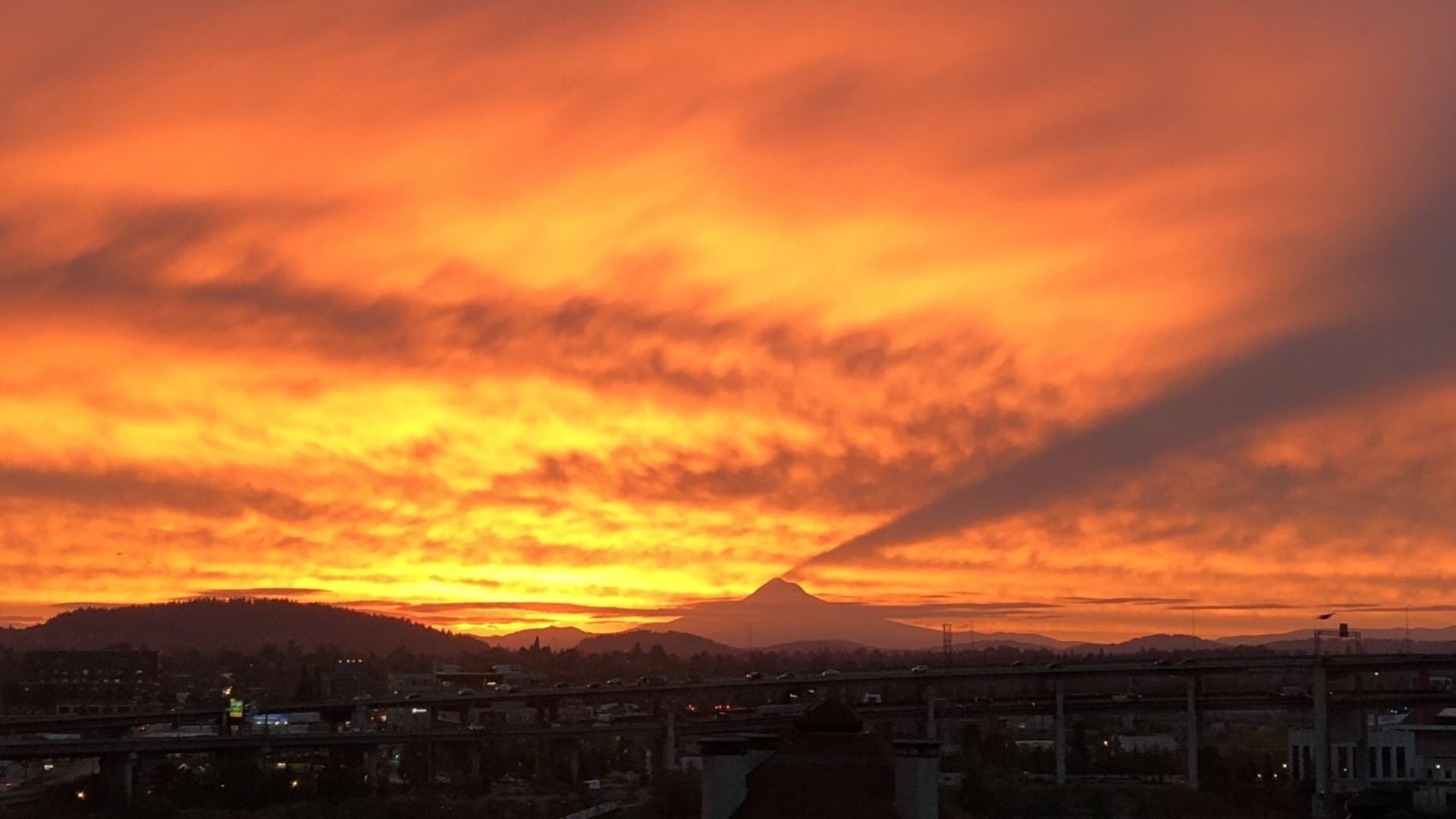 Vivid orange and red sunset paints the sky, with dark city buildings and distant hills along the horizon; orange clouds glow, while a highway and low structures sit in the foreground.