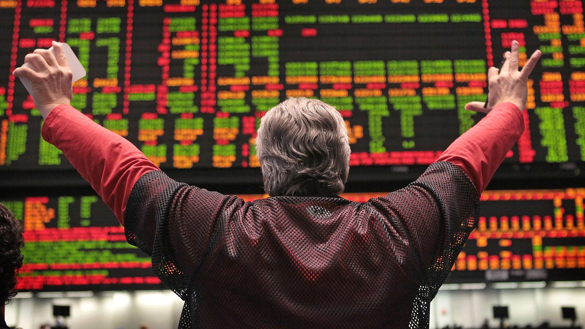 Man with back toward camera with arms in cheering position looking at the stock market ticker