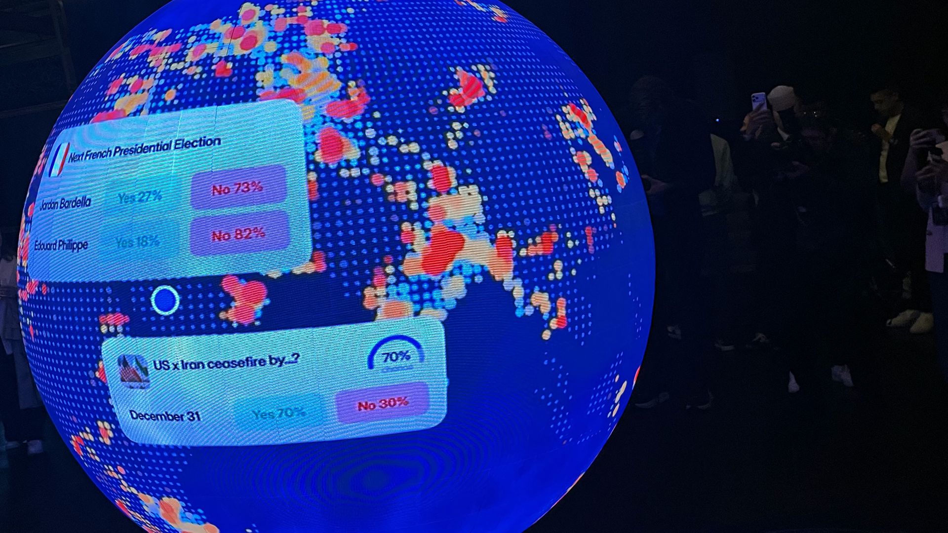 A large blue globe with a dotted world map, lit by LEDs. Screens on the globe show election graphics while a dark room and people taking photos are visible in the background.