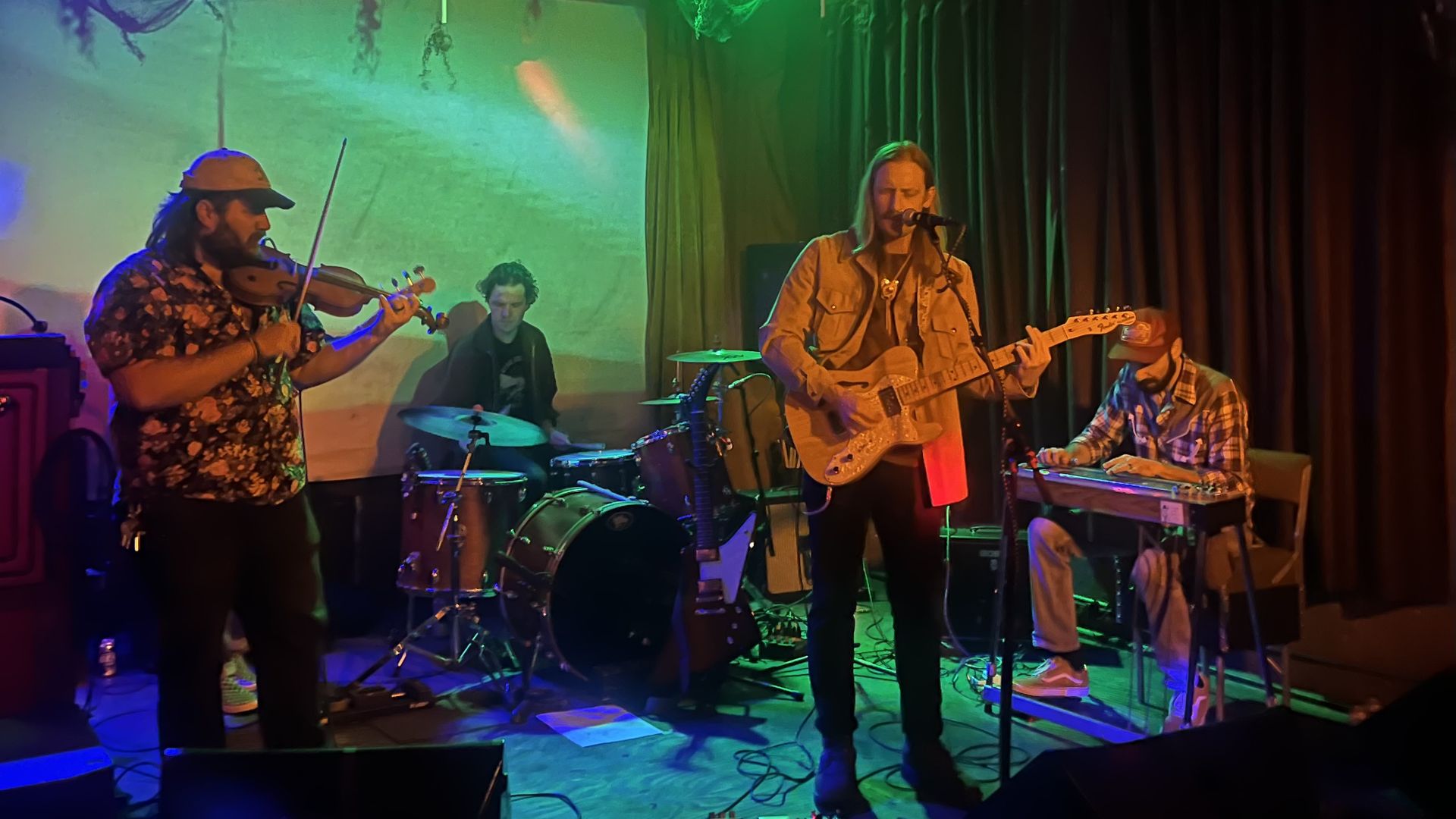 Live band on a dim stage bathed in green and blue lights: left violinist in a floral shirt and cap, center singer-guitarist in a tan jacket, drummer behind, right keyboardist; dark curtains in back.