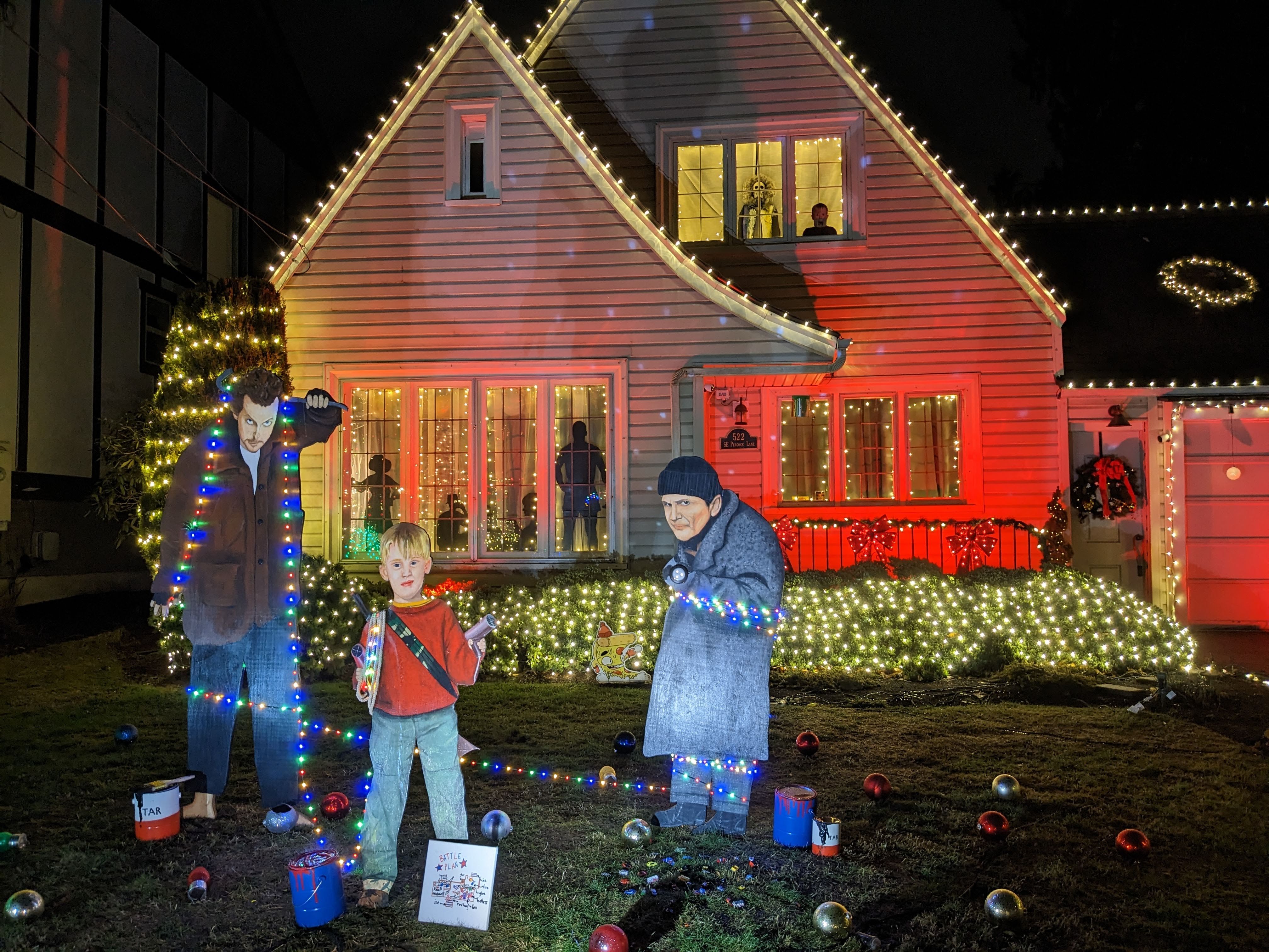 House decorated with white string lights, red bows, and a wreath, featuring cutouts of three characters from "Home Alone" wrapped in multicolored Christmas lights on the front lawn at night.