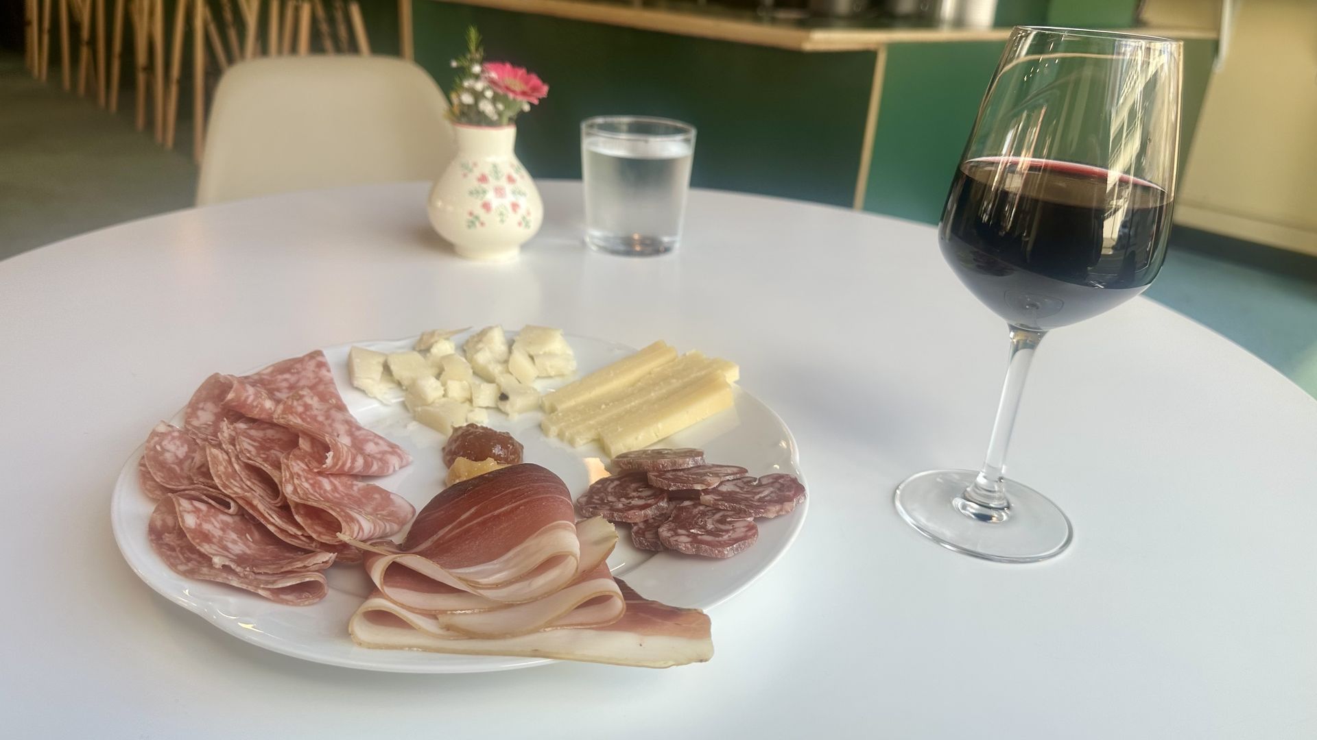 A plate of charcuterie, including three cured meats and two cheeses, on a white table next to a glass of red wine.