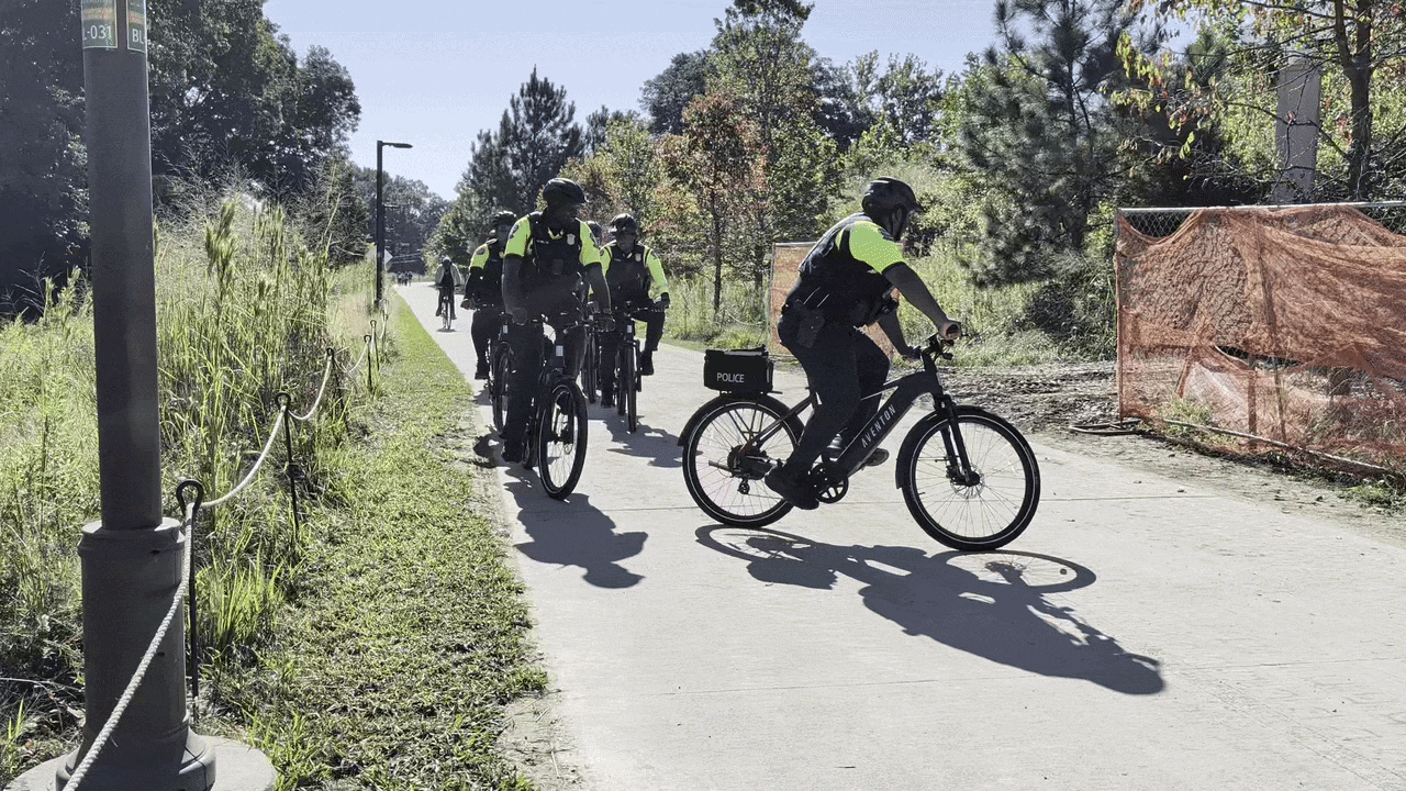 A gif of police officers wearing neon yellow vests riding bikes on a sunlit paved path, lined with tall grass and trees. An officer on a bike with a police bag follows; orange construction fencing is visible on the right.