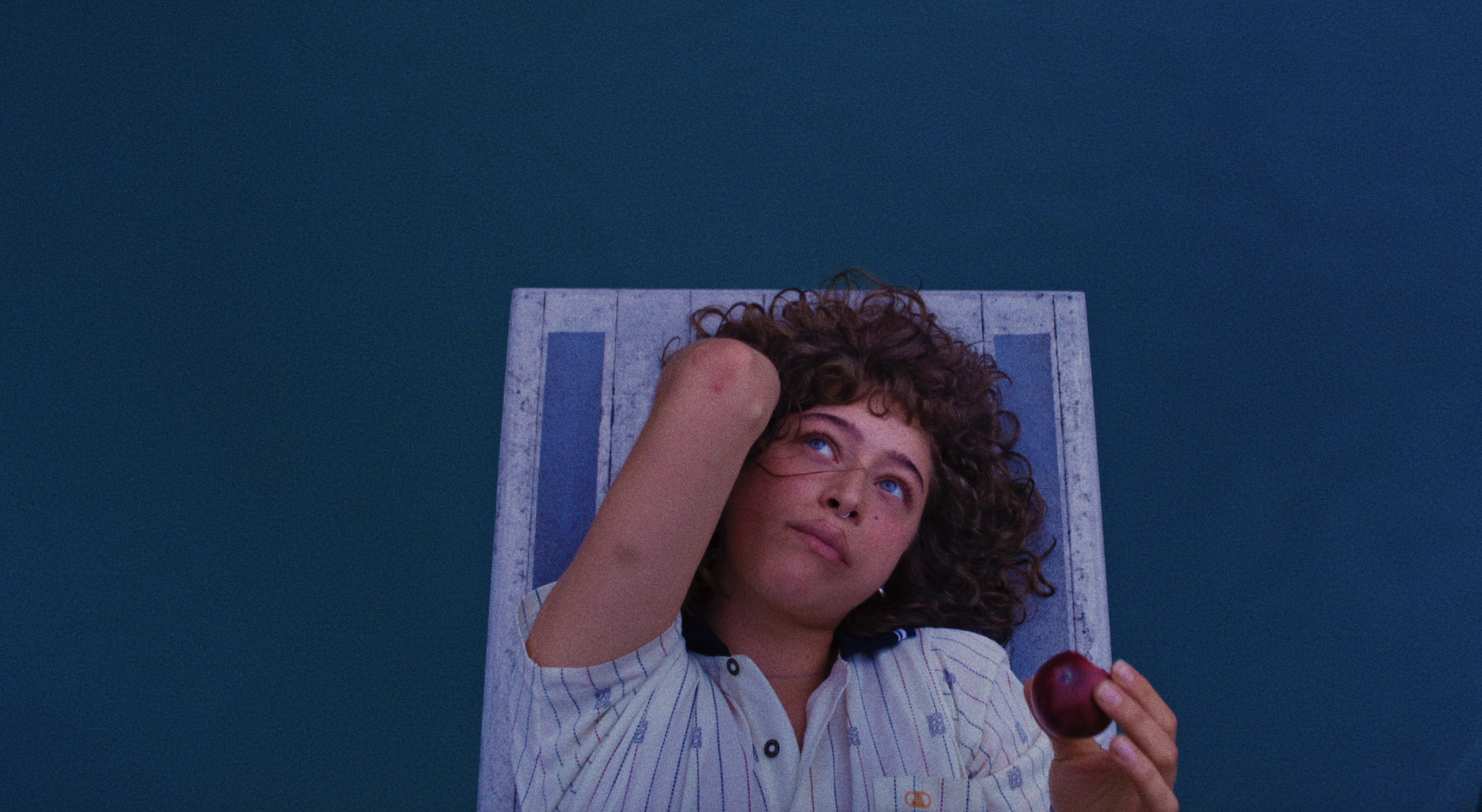 Curly-haired person in a striped shirt lying on a wooden platform above blue water, holding a small red apple while looking thoughtfully upward.