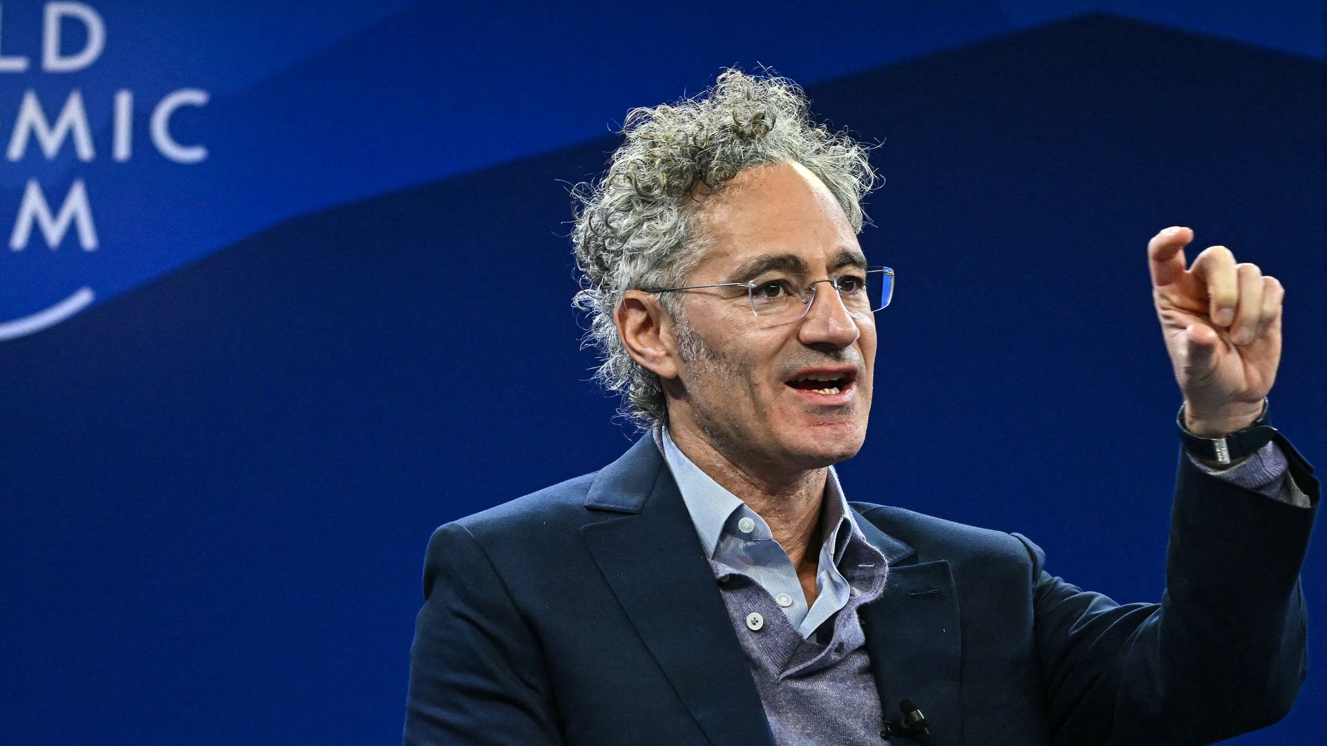 Man with gray curly hair and glasses in a blue suit speaks on stage, raising his hand; a deep blue backdrop displays the logo "World Economic Forum".