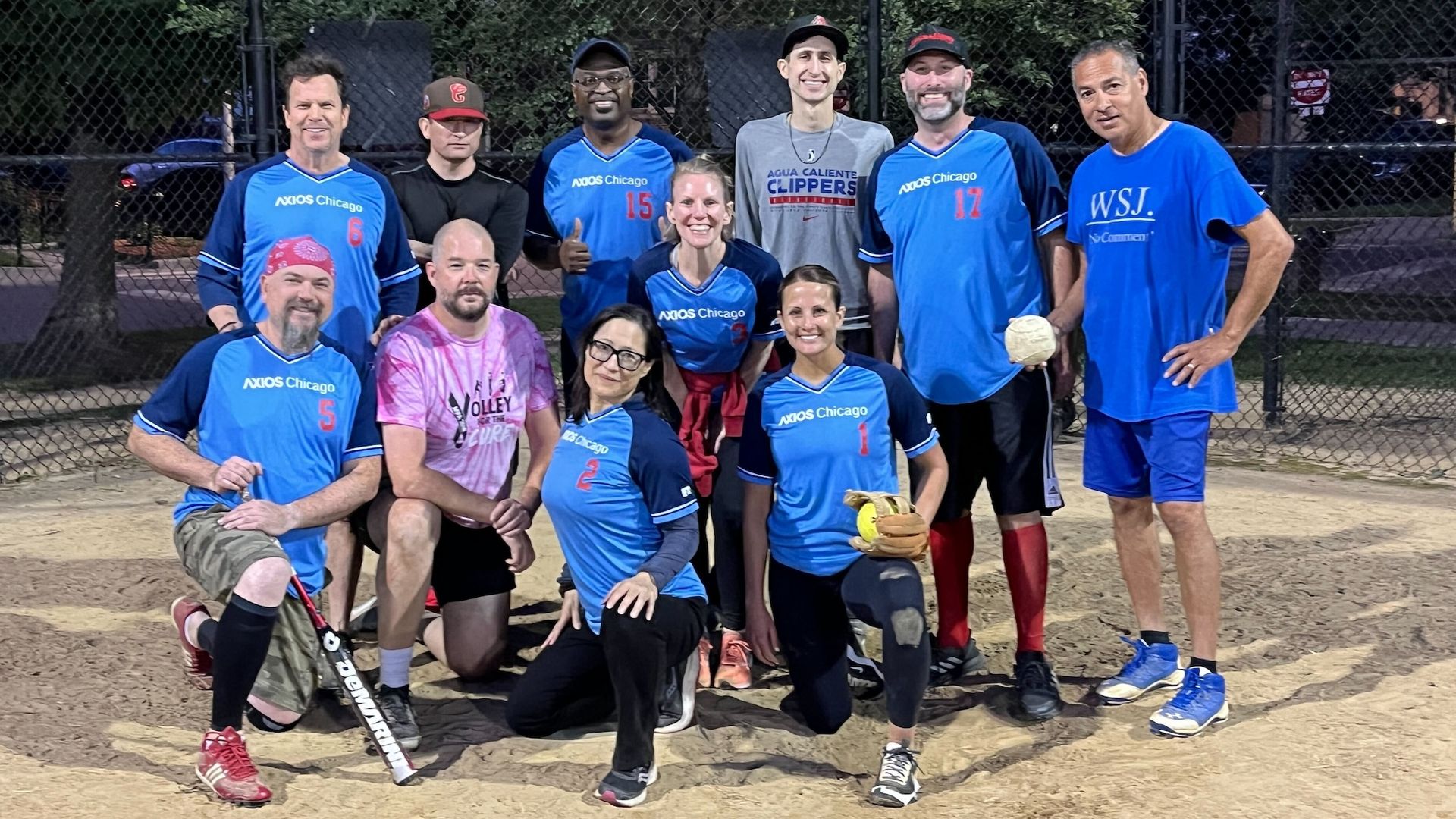 A coed softball team poses at night on a dirt field, wearing blue "AXIOS Chicago" jerseys, one in pink, another in a gray "Agua Caliente CLIPPERS" shirt, and a man in a blue WSJ shirt holding a ball.