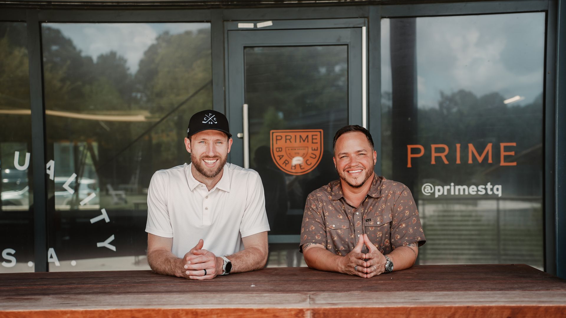 Two men sit at a wooden counter outside a glass storefront with orange PRIME signage; one wears a black cap and light polo, the other a patterned brown shirt, both smiling.