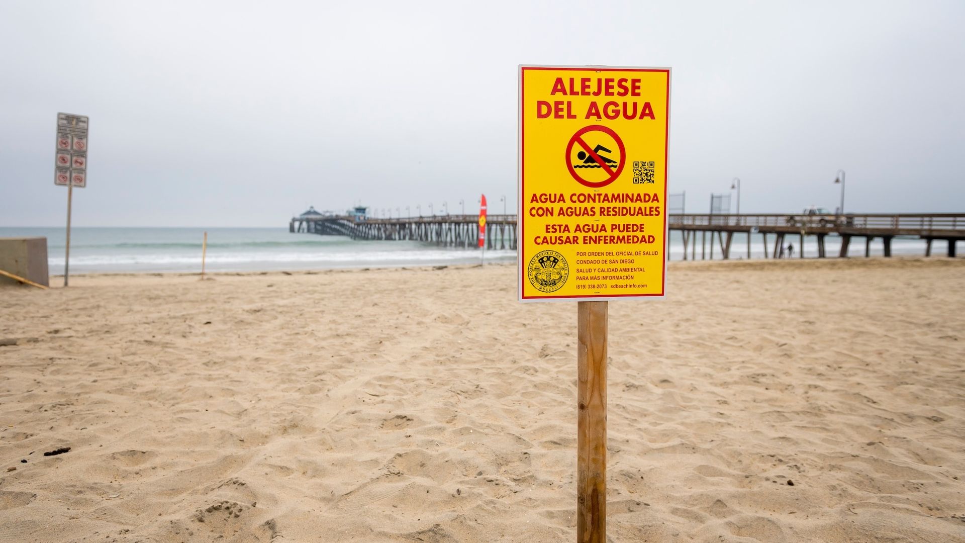 Yellow and red warning sign on sandy beach near pier reading in Spanish to stay away from water due to contamination that may cause illness.