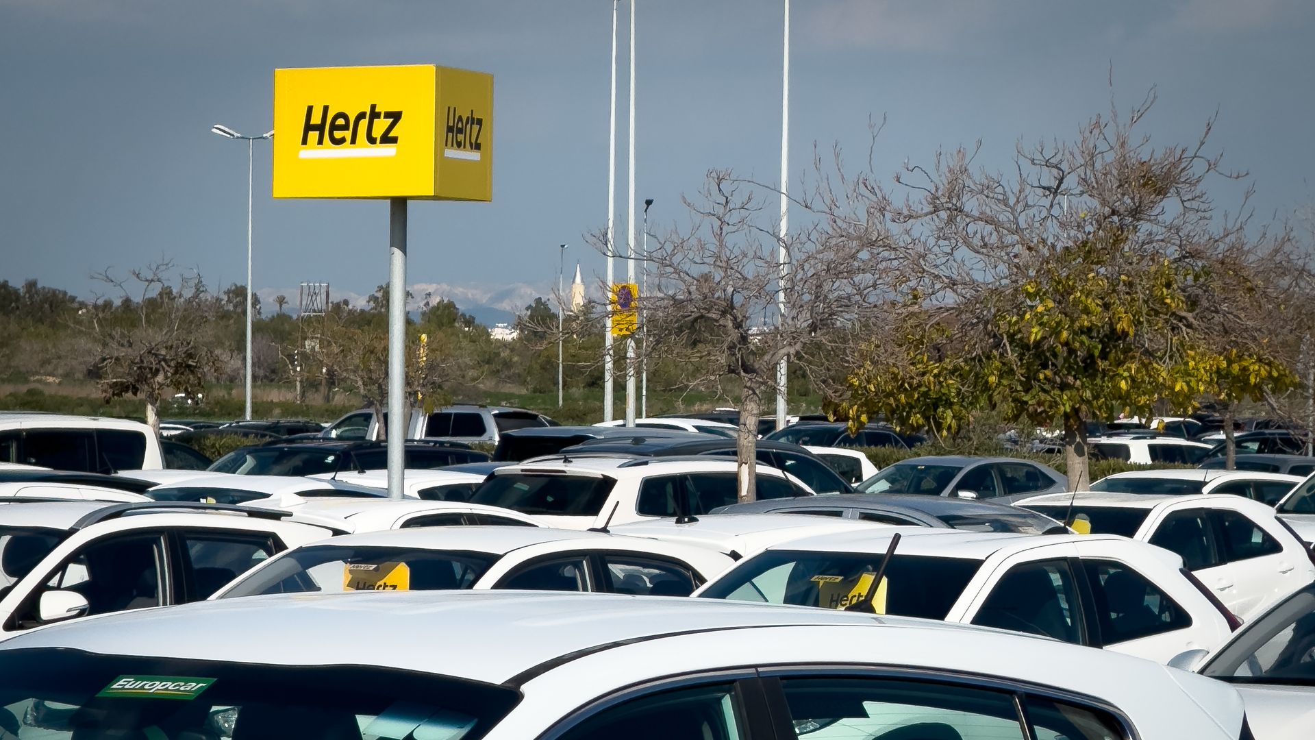 A parking lot full of white rental cars, with a Hertz sign visible in the background.