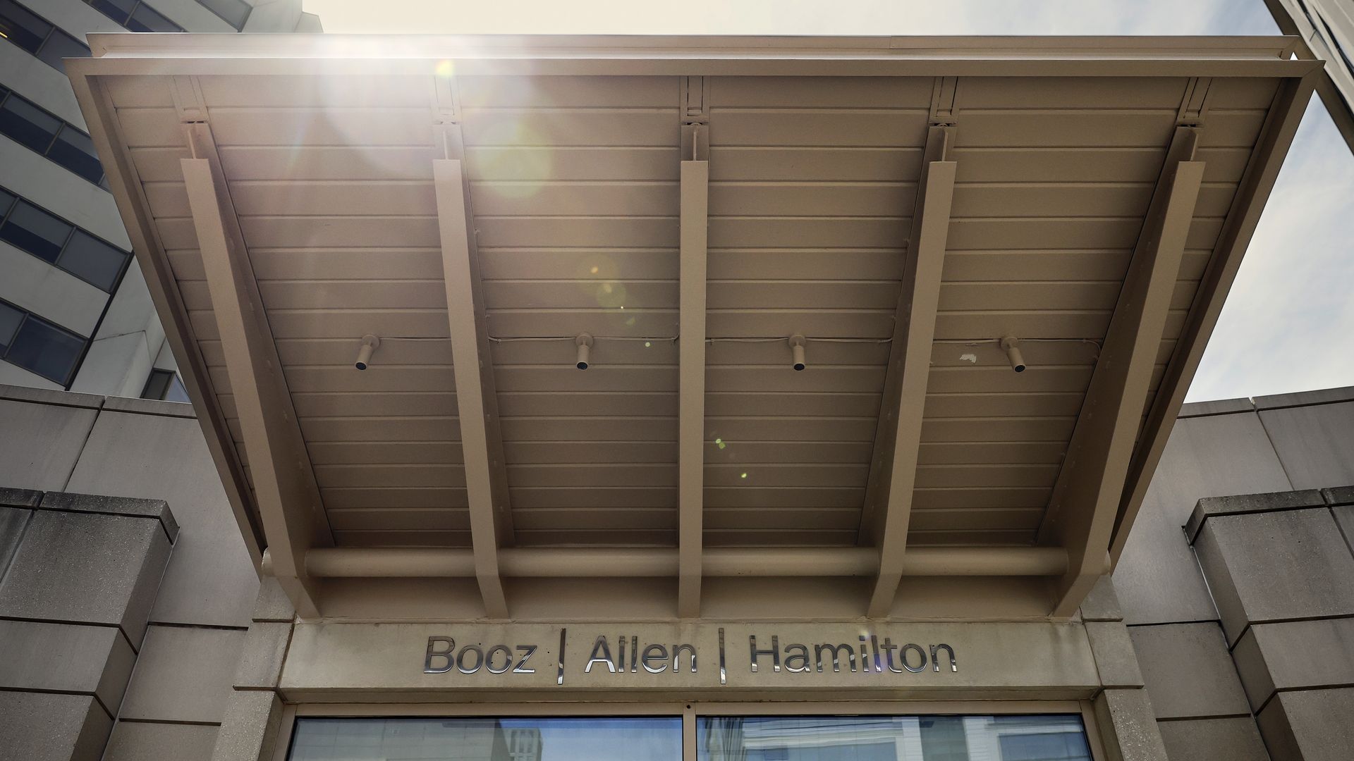Entrance of a building with beige overhead canopy and the name "Booz Allen Hamilton" displayed above glass doors under a partly cloudy sky.